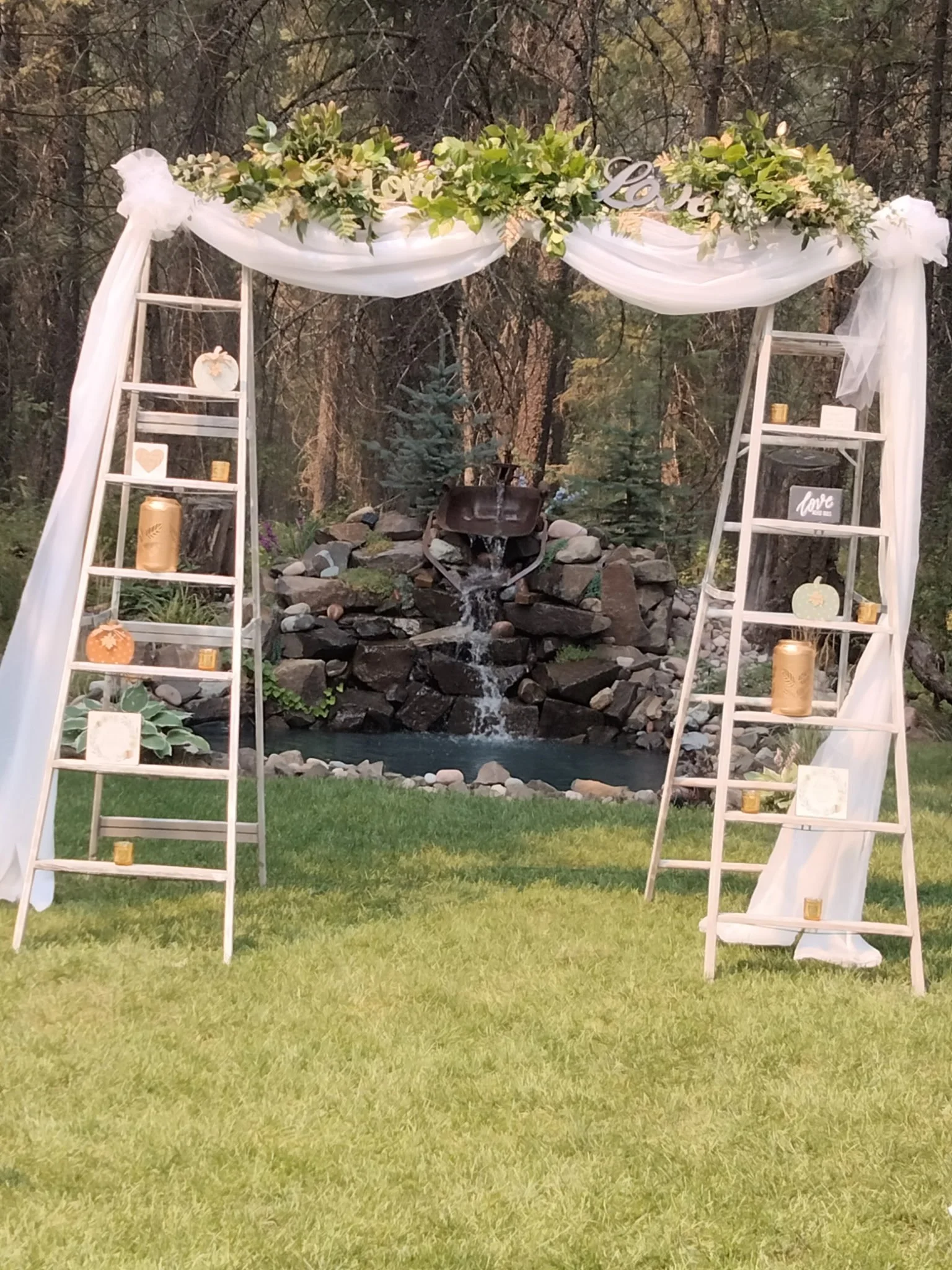 Decorative outdoor wedding arch with white drapery, greenery, and fall-themed accents, set in front of a pond and trees.