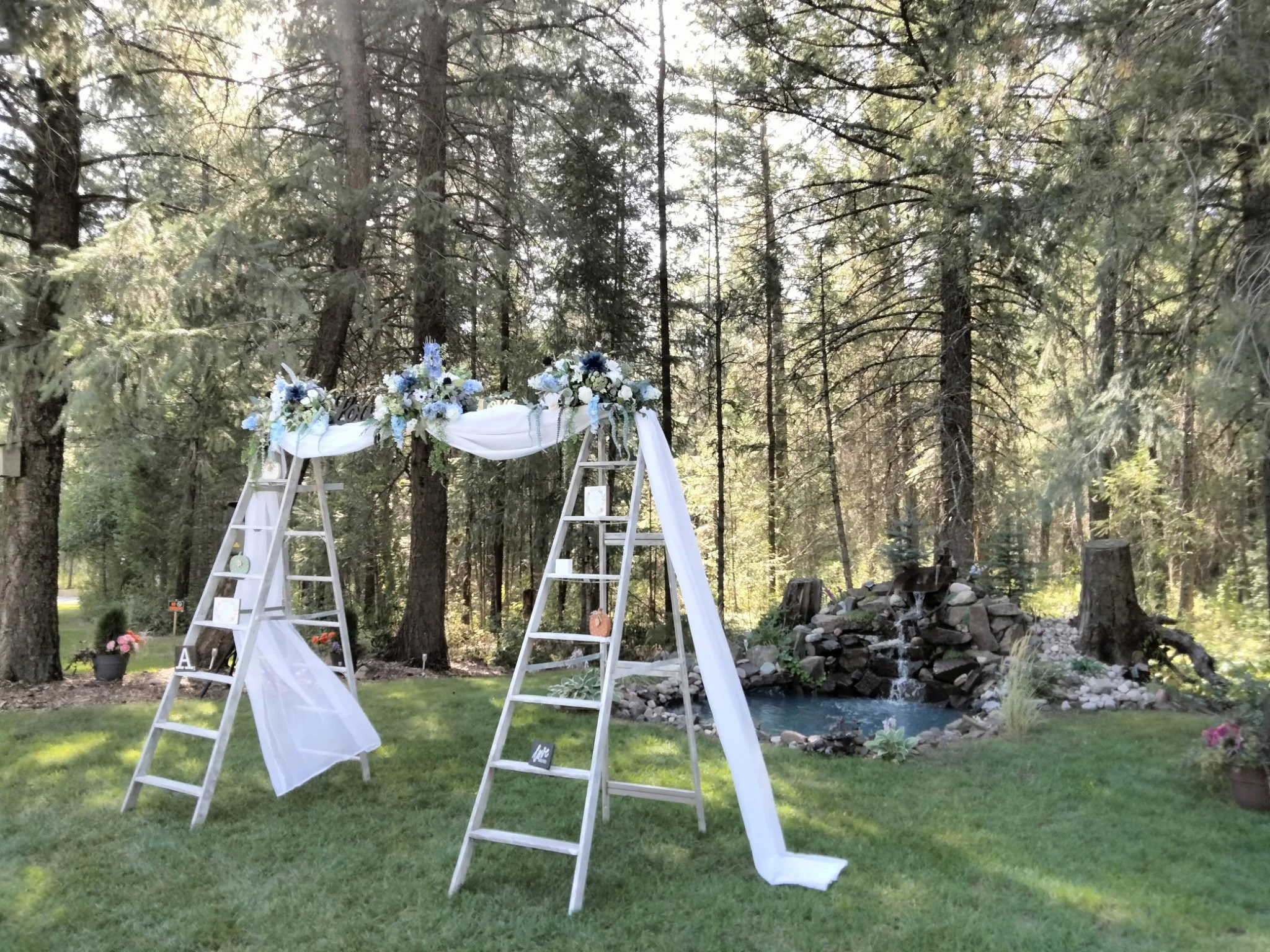 Wedding arch decorated with white and blue flowers, white draped fabric, set outdoors in a wooded area with a small pond and waterfall feature in the background.