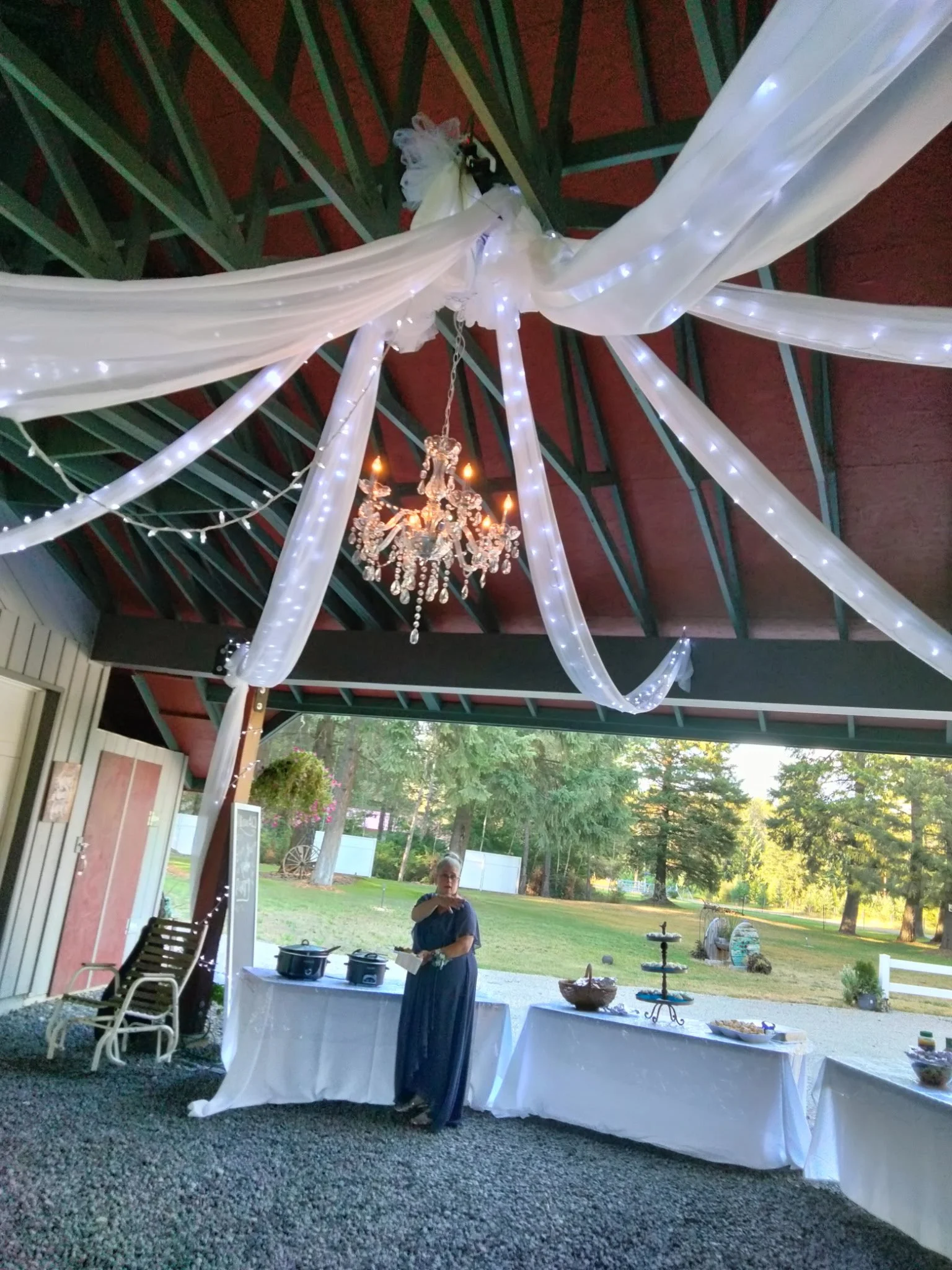 Outdoor event space decorated with white fabric and string lights hanging from a ceiling with a chandelier. A woman stands near tables with food and decorations in the background, with trees and greenery outside.