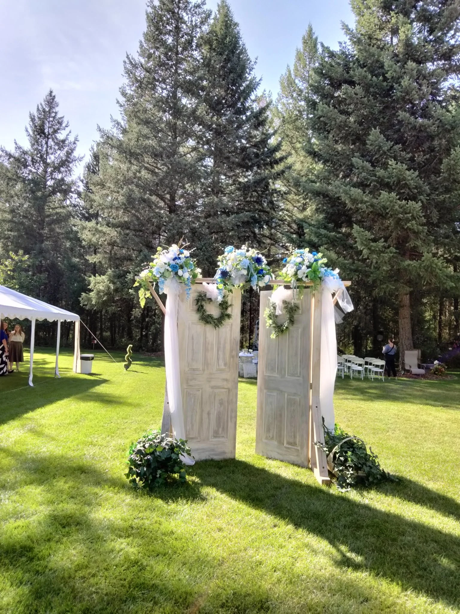 An outdoor wedding arch made of four wooden panels decorated with white and blue flowers and greenery, set on a grassy area surrounded by tall pine trees under a clear sky.
