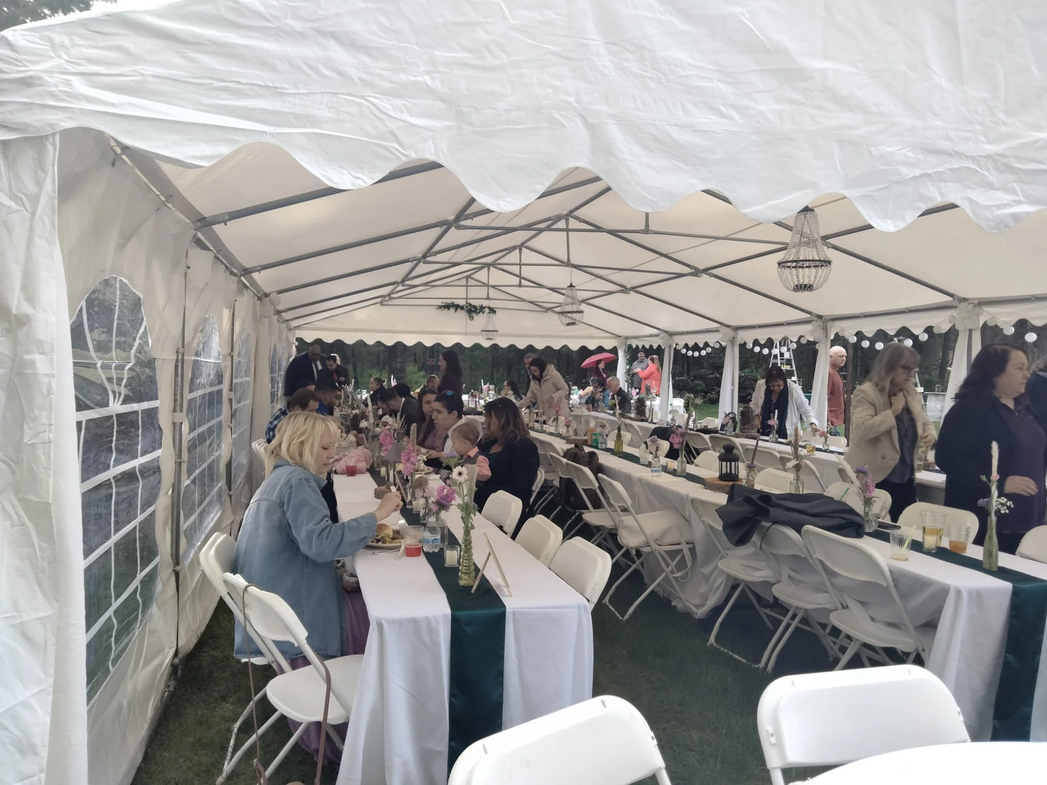 People attending a social event inside a large white tent with long tables decorated with flower vases and lanterns, some people are seated eating, while others are standing and walking around.