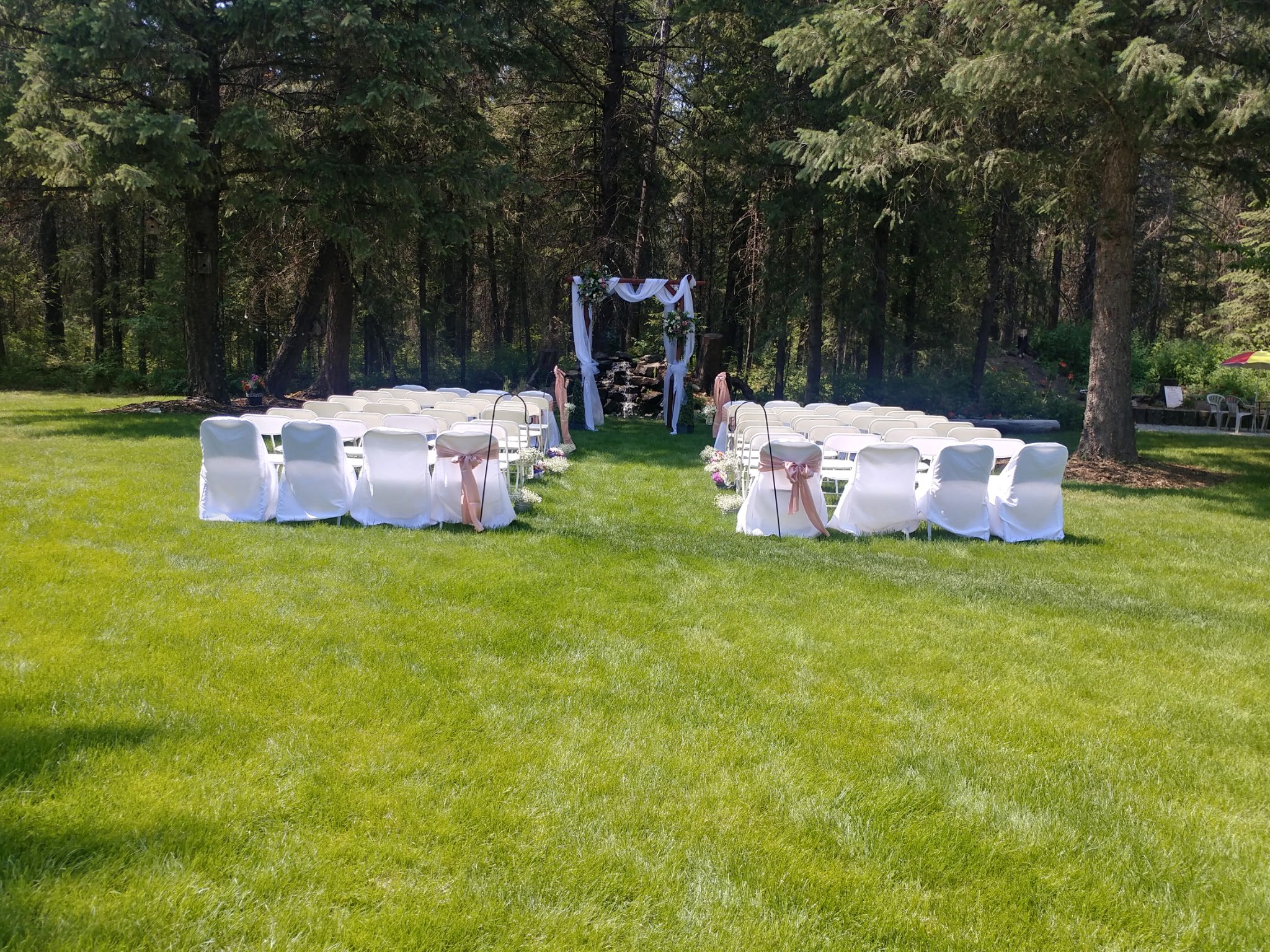 Outdoor wedding ceremony setup with white chairs covered in white cloth and decorated with pink ribbons, arranged in front of a wooden arch draped with white fabric and flowers, on a lush green lawn surrounded by tall trees.