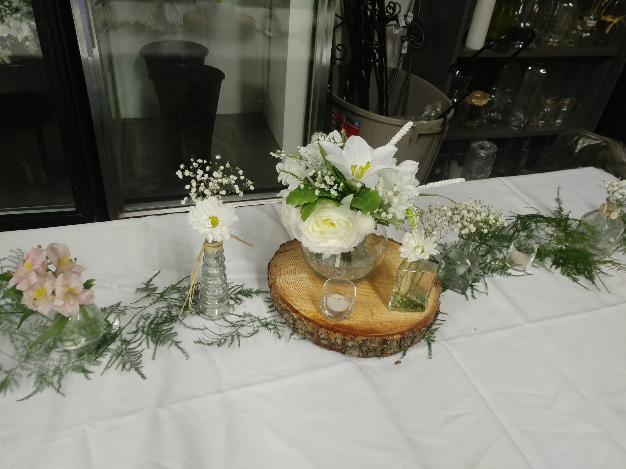 Decorative table centerpiece with white flowers, leaves, and candles on a wooden slab, with additional floral arrangements along a white cloth table.