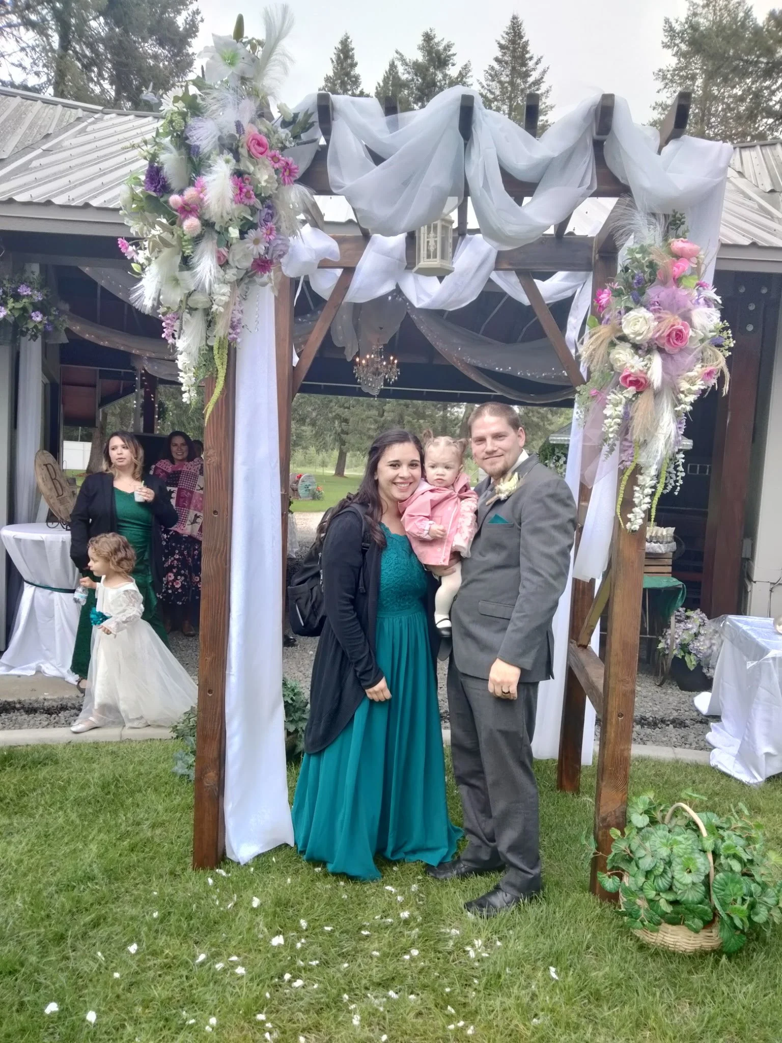 A family at a wedding, standing under a decorated wooden archway with white fabric and pink and white floral arrangements. The woman is wearing a teal dress, the man is in a gray suit, and they are holding a small child. In the background, other gues