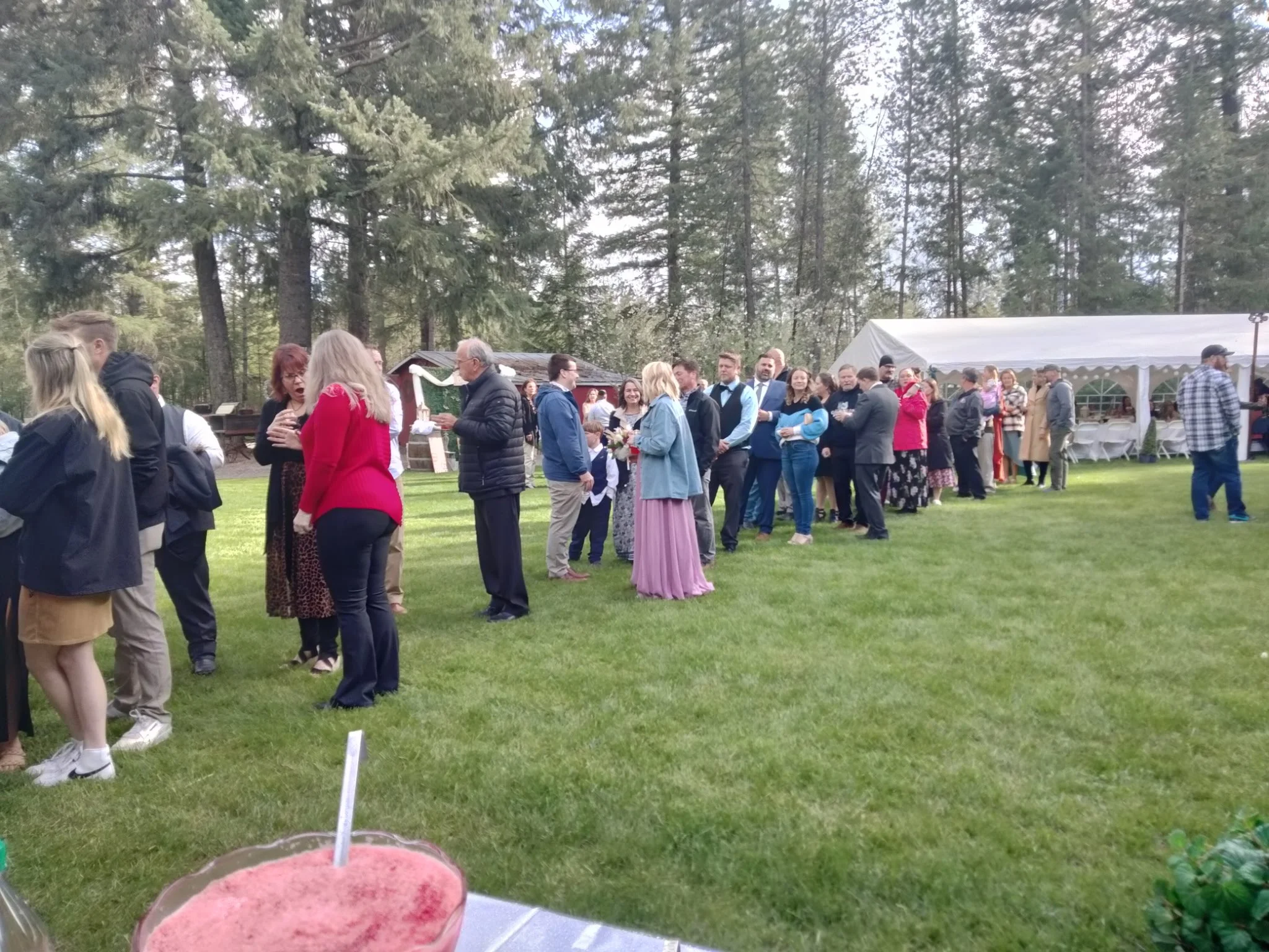 People standing in a line outdoors at a gathering or event, with a white tent and trees in the background.