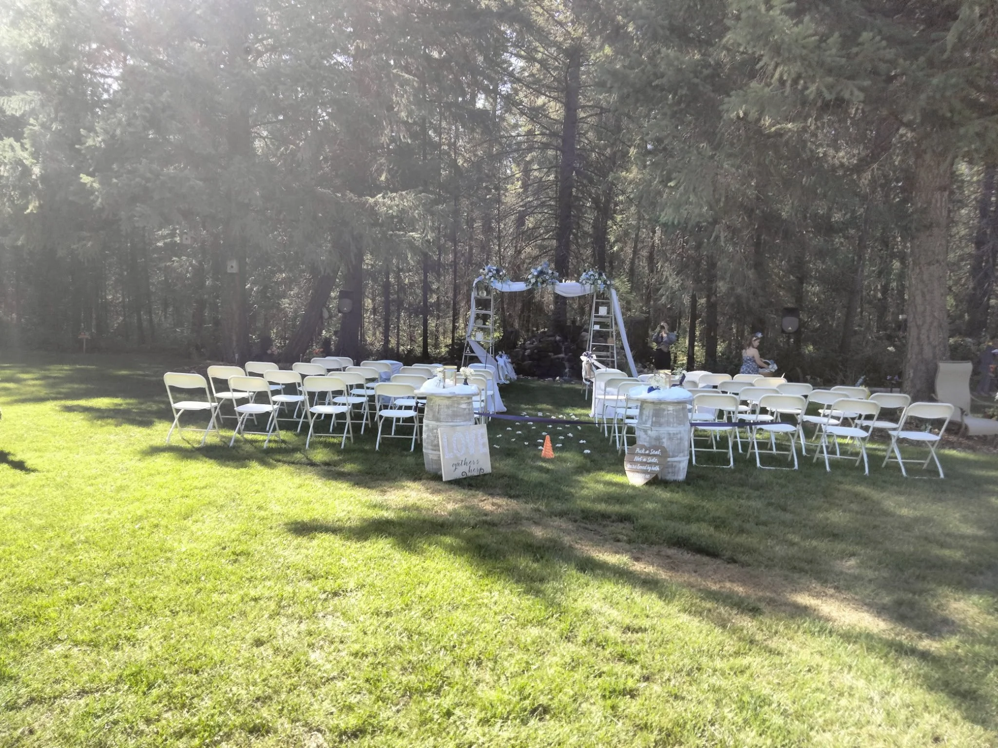 Outdoor wedding ceremony setup on grassy field with white chairs arranged in two sections facing a gazebo decorated with flowers and white fabric, surrounded by tall trees.