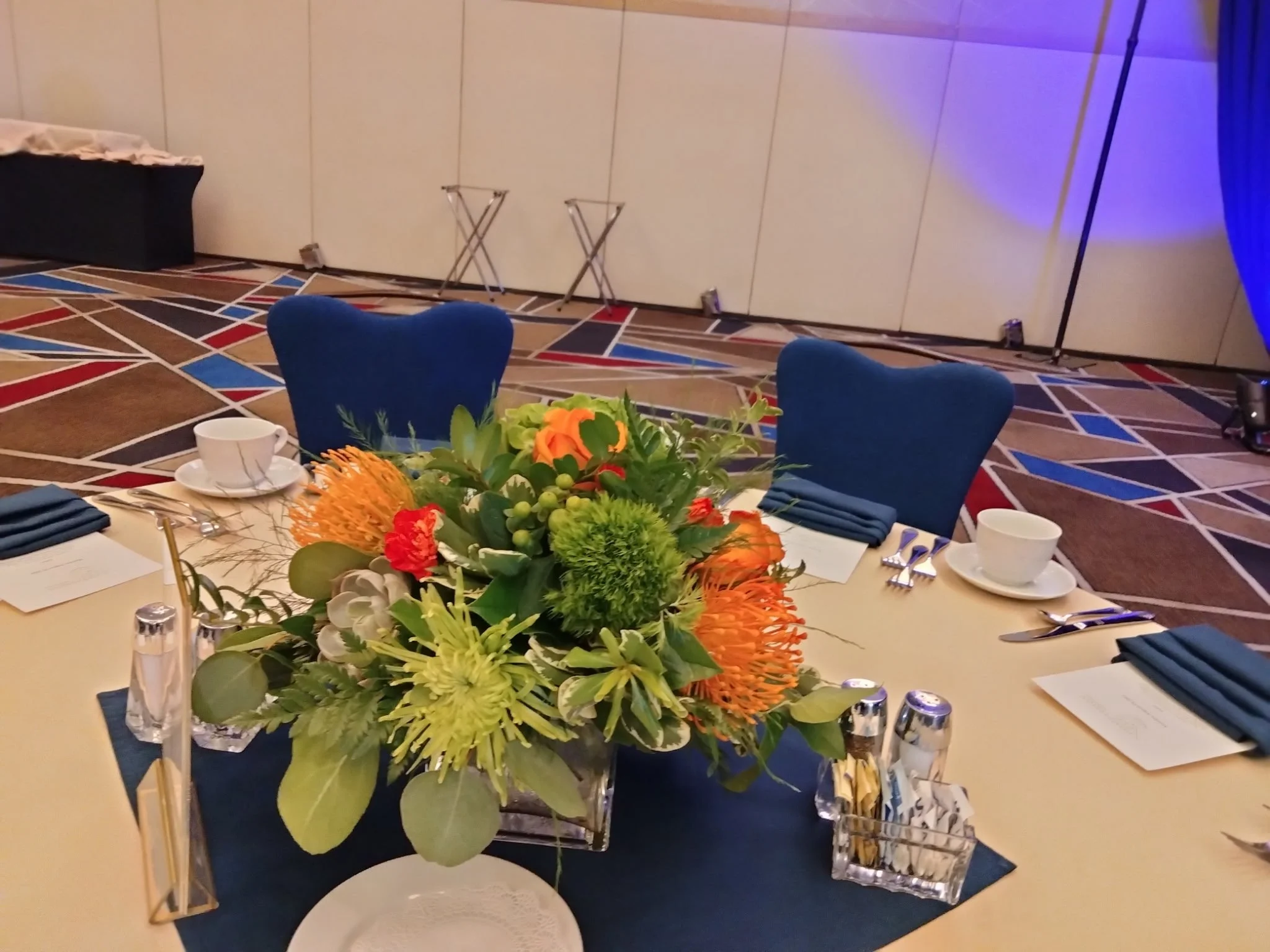 Round table decorated with a large flower arrangement, set with blue napkins, plates, cups, utensils, and water glasses in a banquet hall.