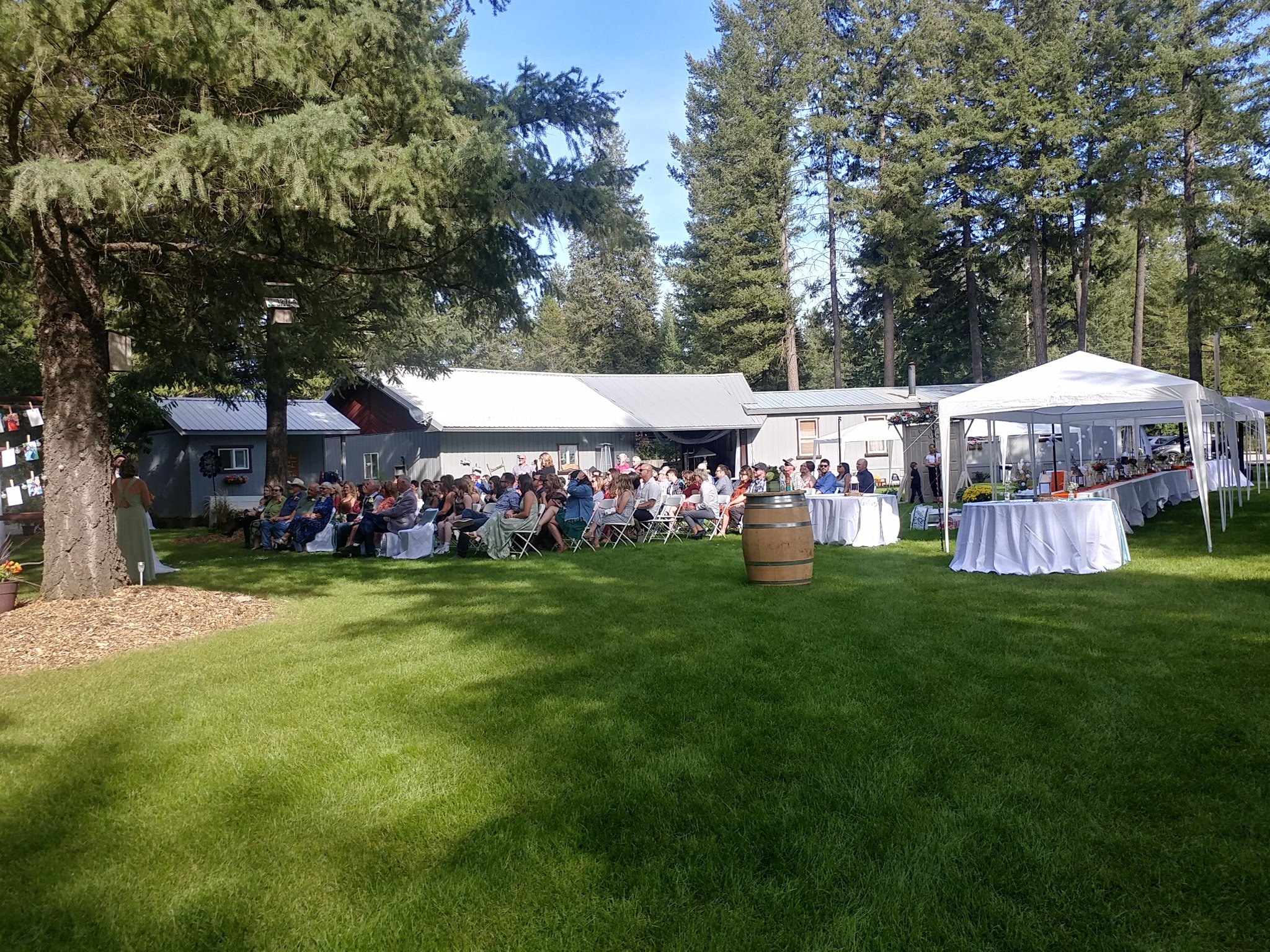 Outdoor wedding reception with guests seated in chairs and lawn, white tents with buffet tables, large trees, and a rustic building in the background on a sunny day.