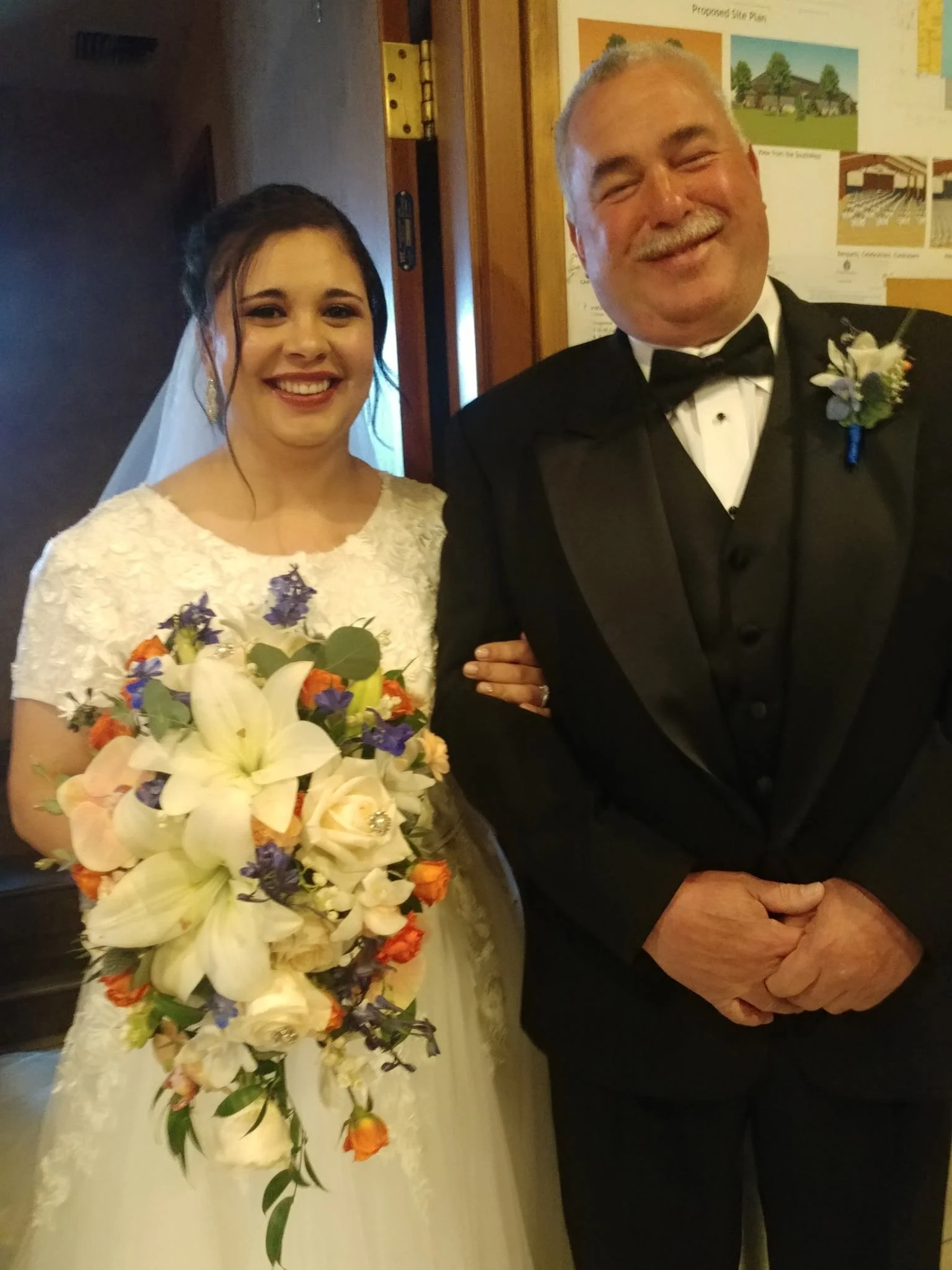 A bride and an older man, possibly her father, smiling at a wedding. The bride wears a white lace wedding dress and holds a large colorful bouquet, while the man wears a black tuxedo with a boutonniere. They are indoors, with wedding posters visible 