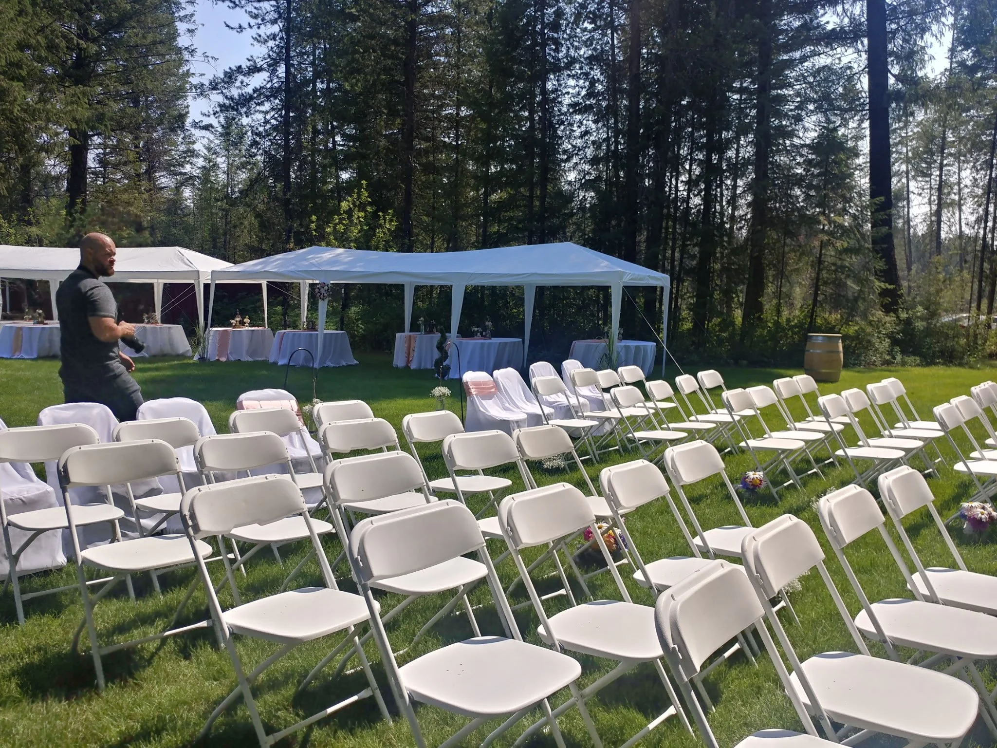 White folding chairs arranged outdoors on green grass for a wedding ceremony, with tents and tables set up in the background surrounded by trees on a sunny day.