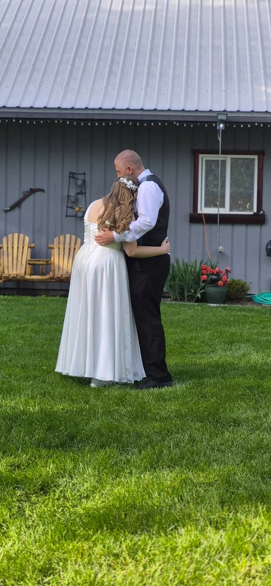 A bride and groom embracing in a yard with grass, next to a gray building with a window and flowerpots, during a wedding celebration.