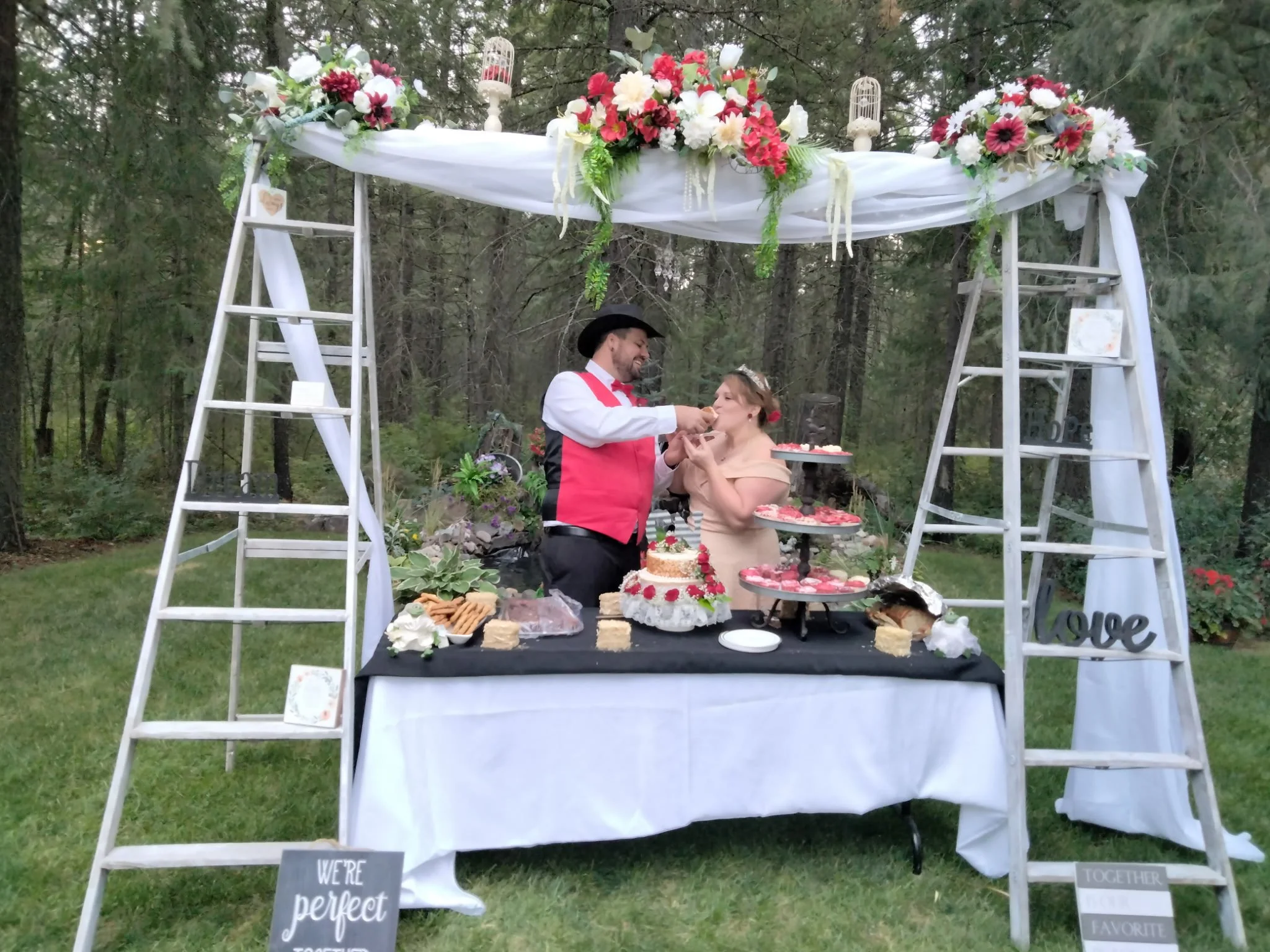 A couple at an outdoor wedding reception standing behind a table with birthday cake, cookies, and desserts, under a decorated canopy with flowers and lanterns, in a forested area.