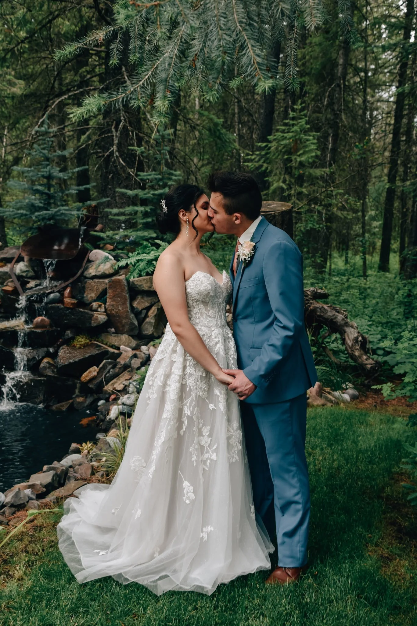 A bride and groom in wedding attire kiss in a lush, green forest setting by a small pond and waterfall.