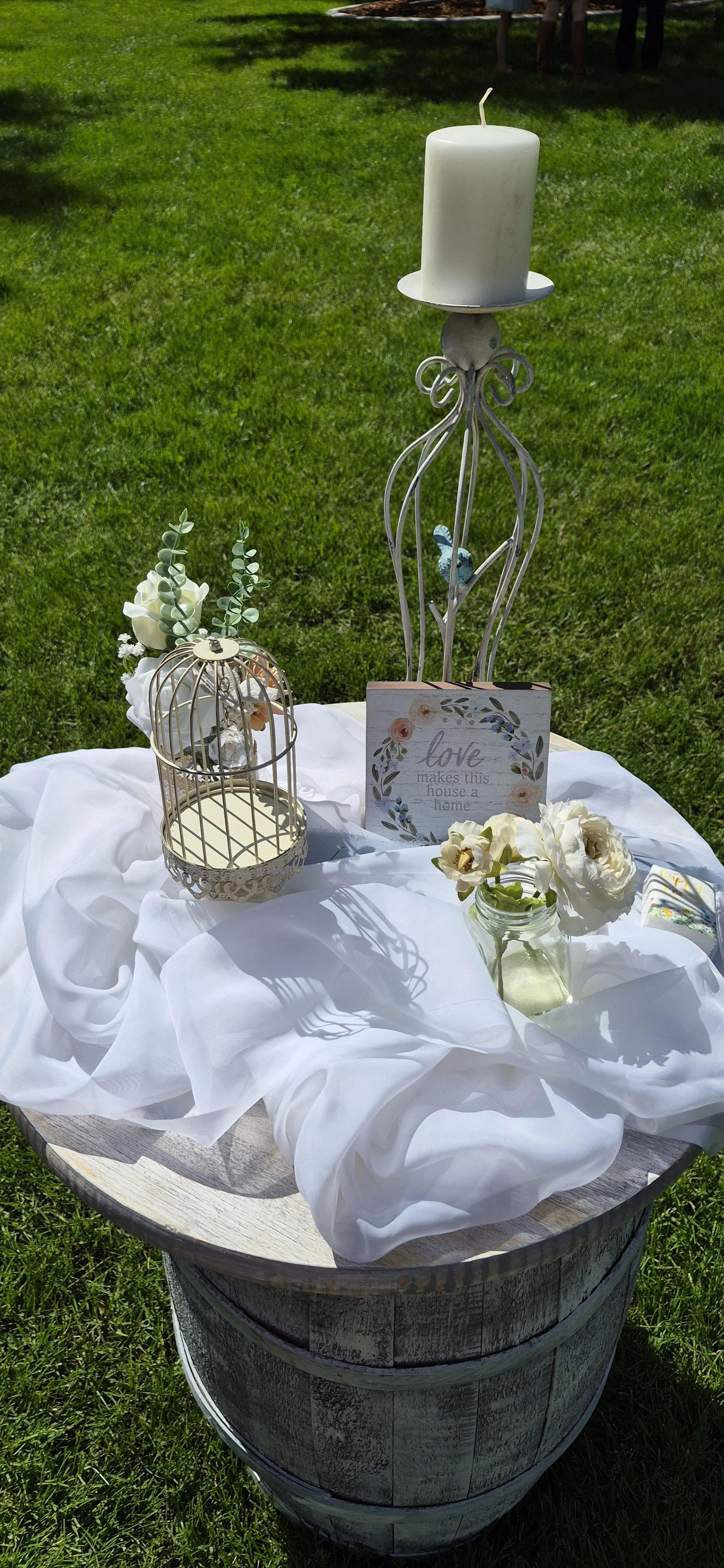 Decorative outdoor table with white cloth, holding a white pillar candle on a metal stand, a small birdcage with flowers, a jar with white roses, and a sign with floral design and the message 'love makes this house a home,' set on grass.
