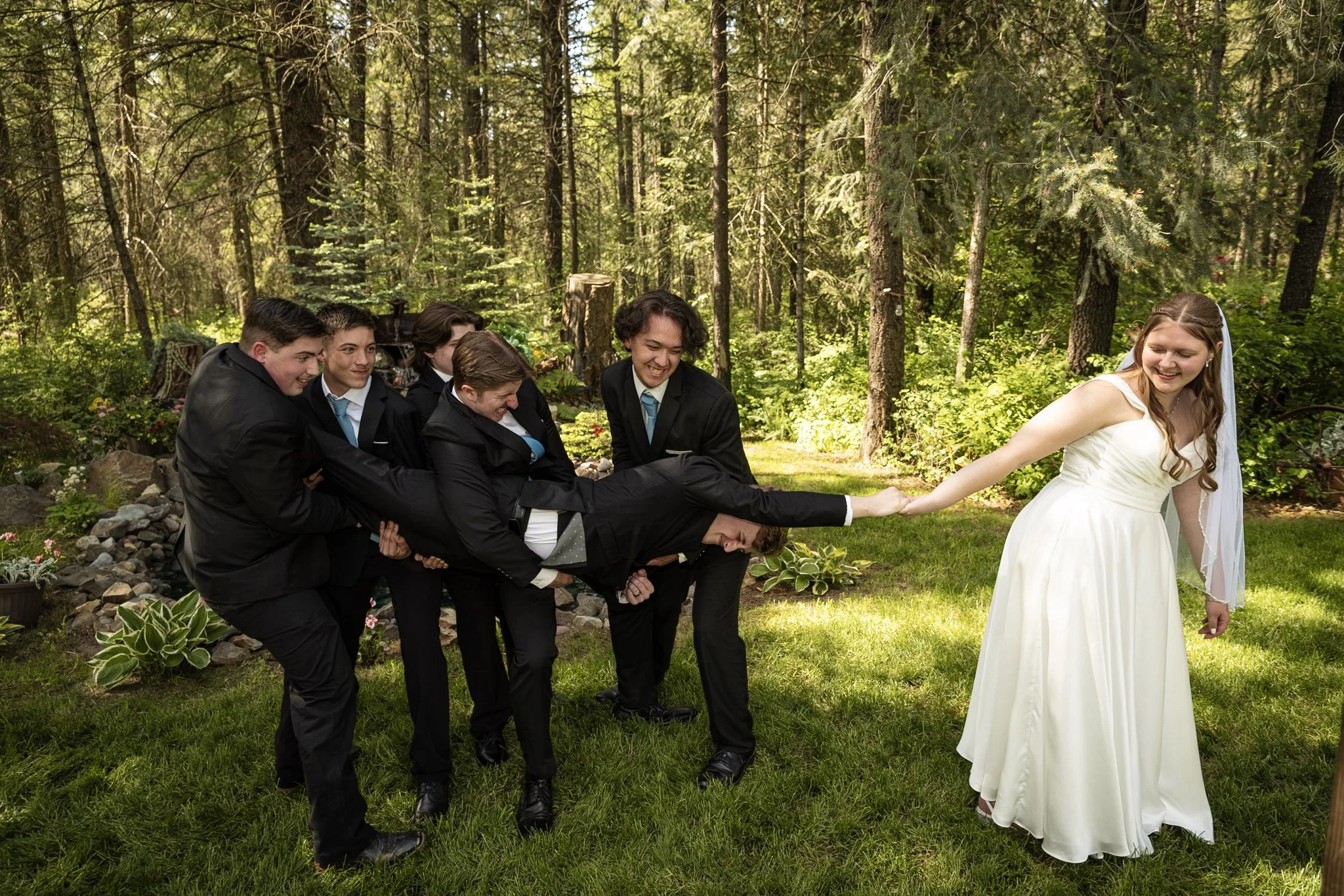 A group of young men in suits playfully holding another young man horizontally while a young woman in a white wedding dress smiles and reaches out her hand towards him in a forested outdoor setting.