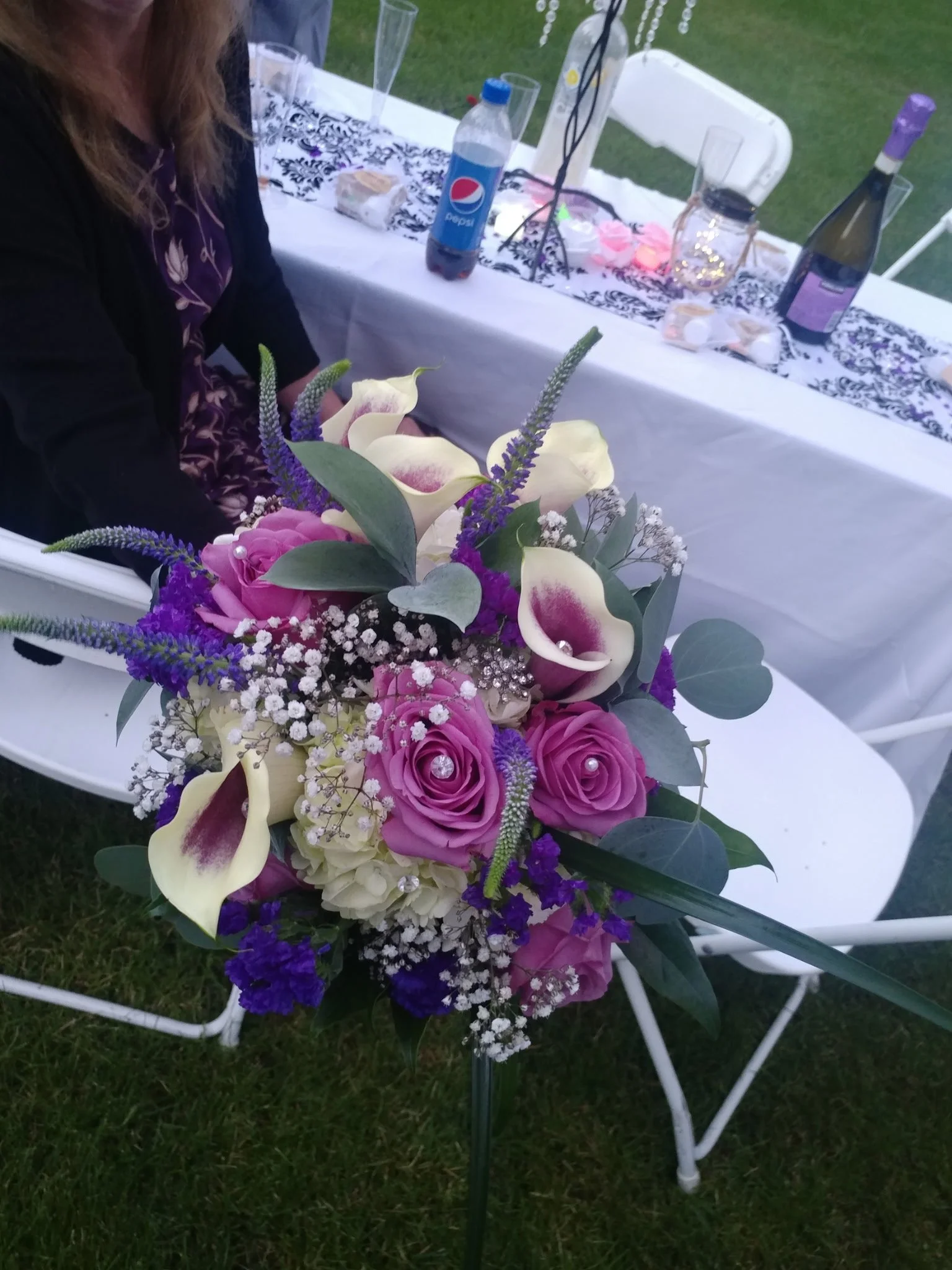 A floral arrangement with pink roses, white calla lilies, purple statice, and other greenery on a garden party table.