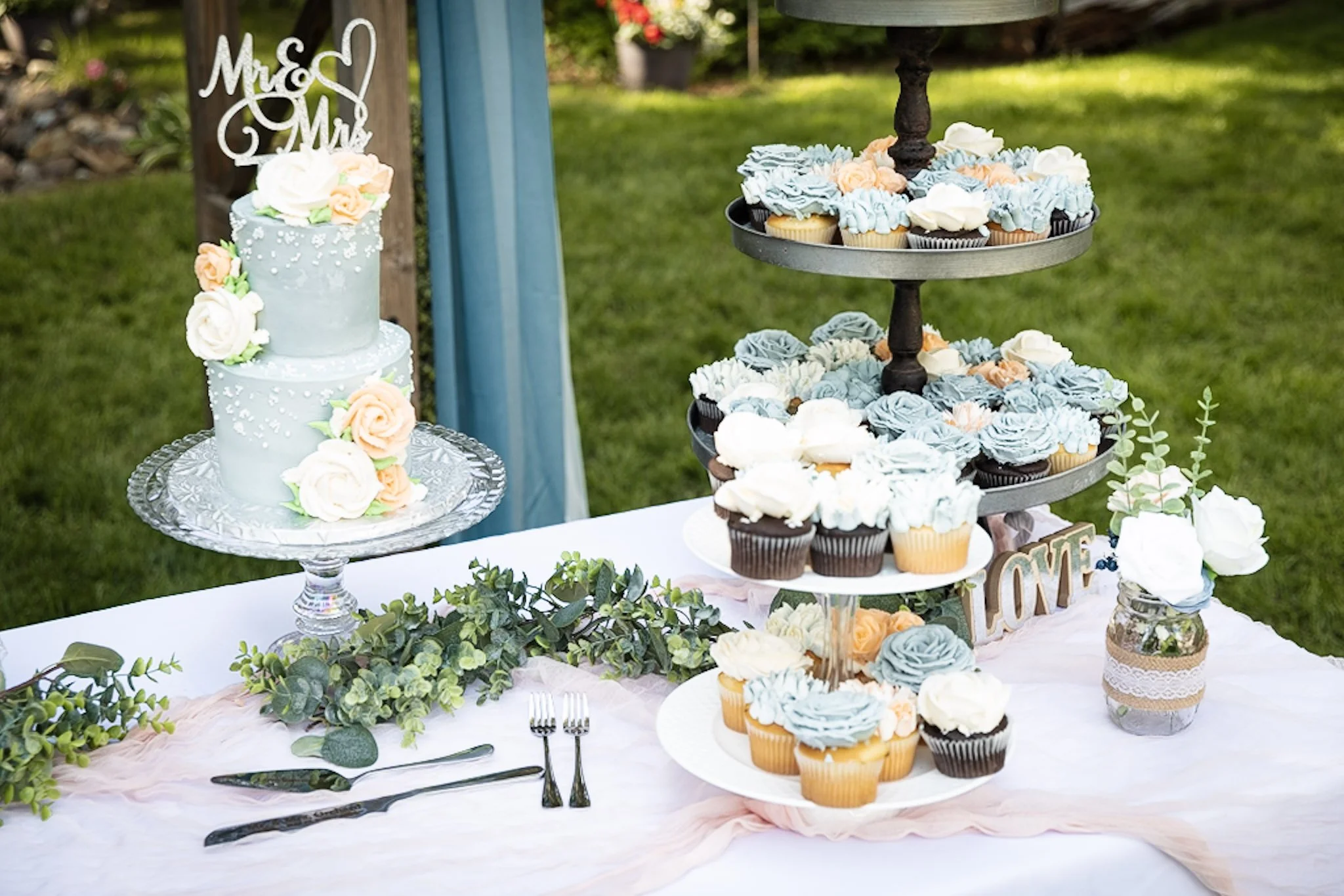 Wedding dessert table with cupcakes decorated with roses and a cake with a wedding topper reading 'Mr & Mrs' in an outdoor garden setting