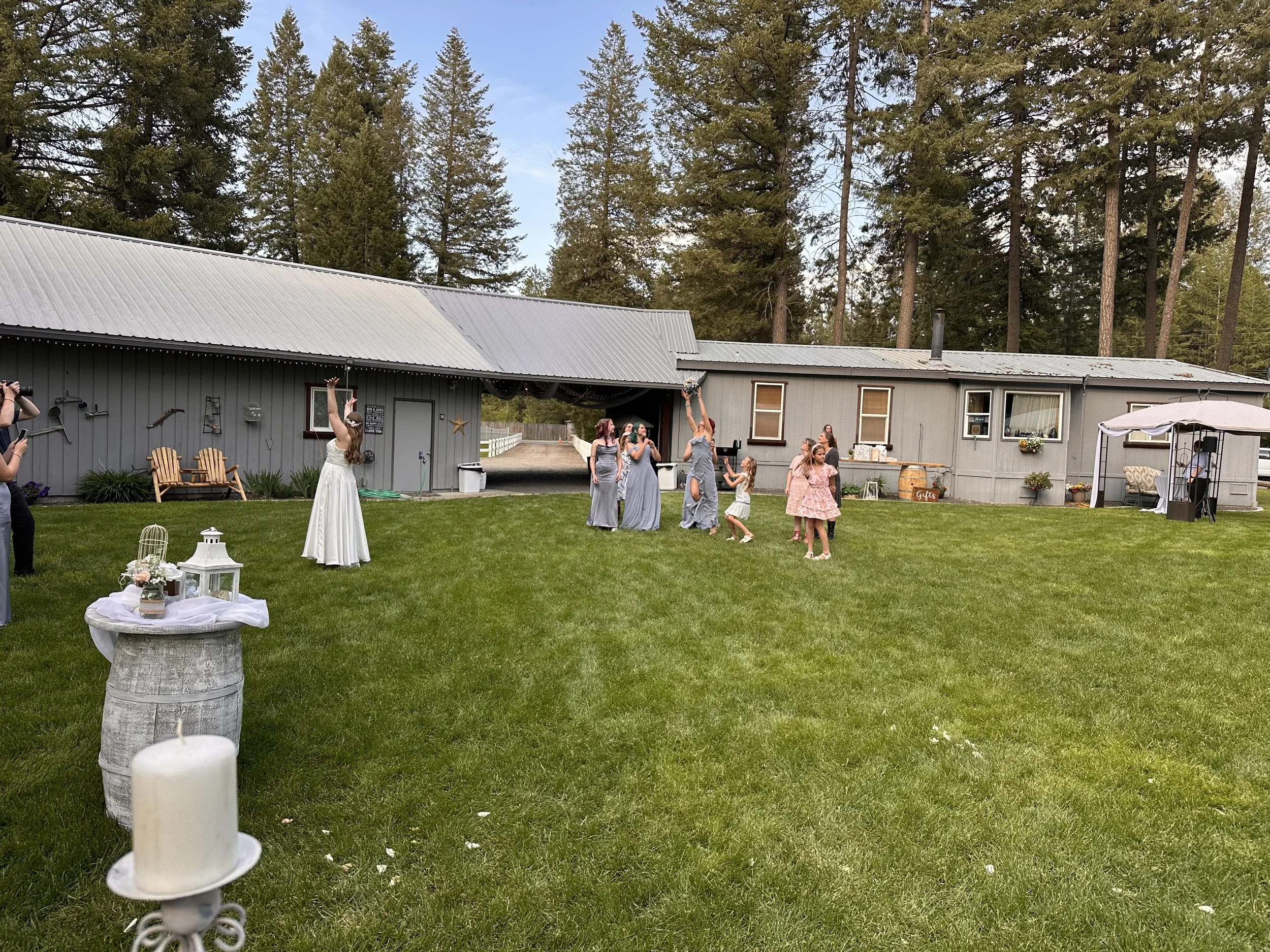 A group of women in dresses celebrating outdoors, possibly at a wedding, on a grassy lawn with a gray building and tall evergreen trees in the background.