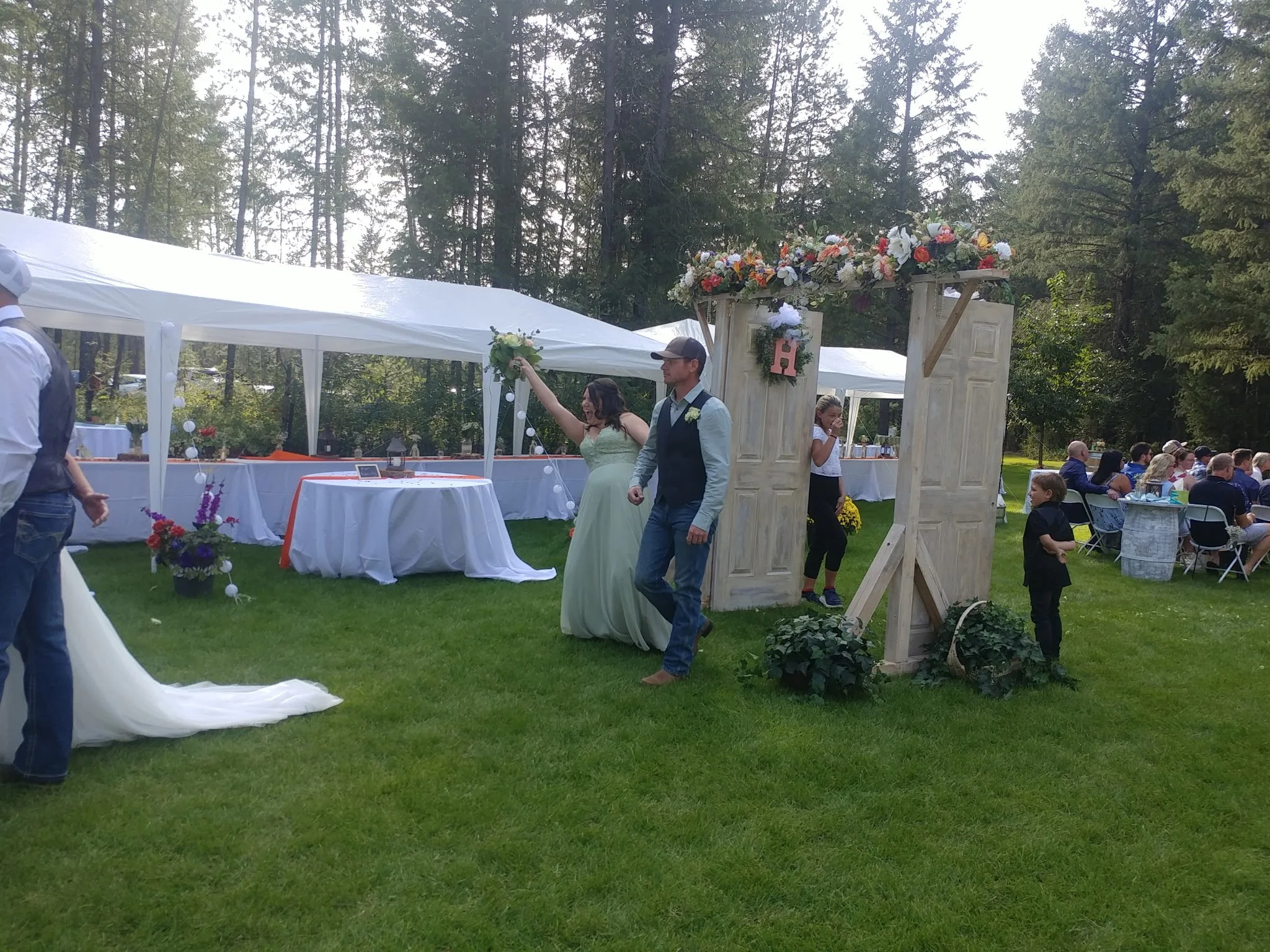 A wedding reception outdoors on a grassy area surrounded by trees. Guests are seated at tables in the background. A bride in a white dress and a groom in a vest and jeans are walking through a decorated archway with flowers. There are white tents wit