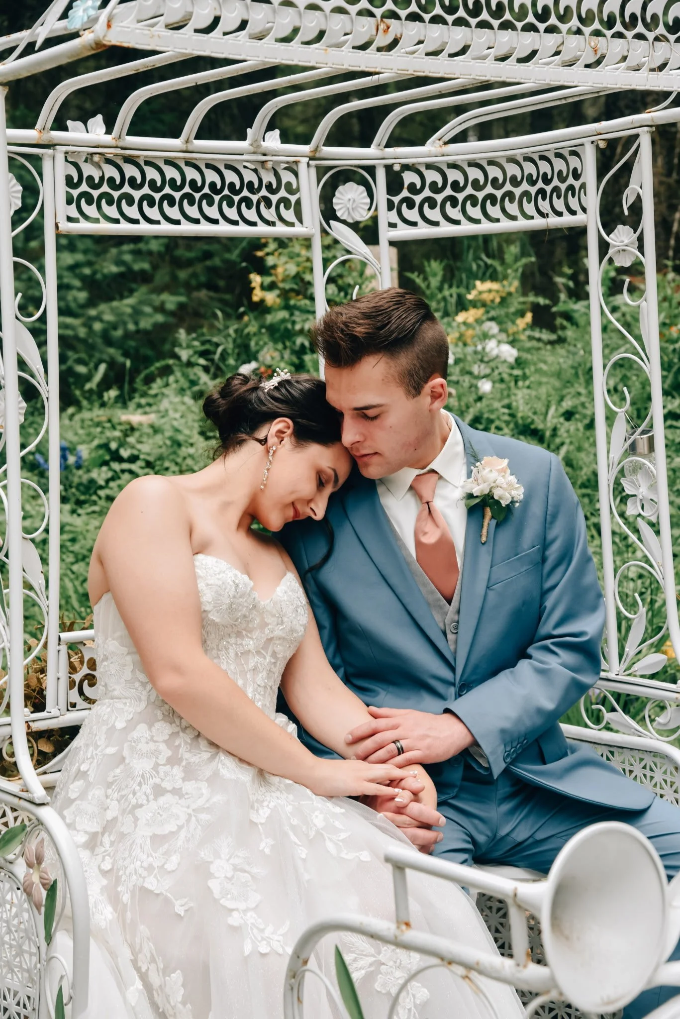 Bride and groom sitting closely on a white metal garden bench, their foreheads touching, with greenery in the background.