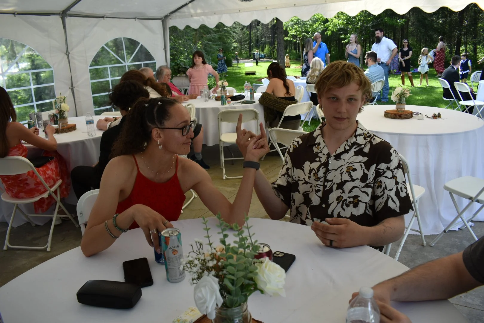 A woman raising her hand and pointing her finger, sitting at a table with a young man at a social gathering or outdoor event. The table has a floral centerpiece, a can of sparkling water, two smartphones, and a water bottle. In the background, people