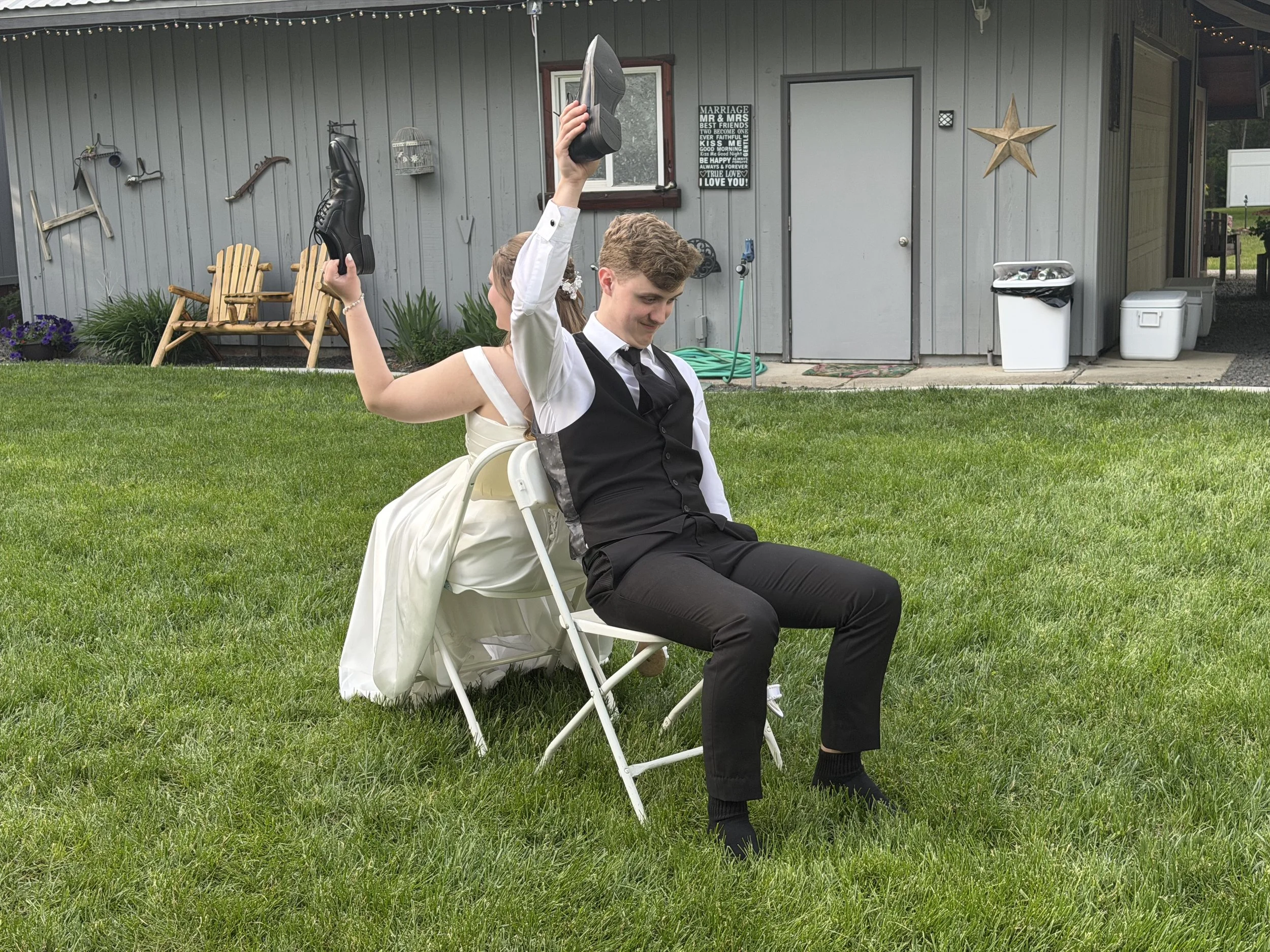A man and woman sitting back to back outdoors in a yard, with the woman young and dressed in a wedding gown, and the man young and dressed in a black suit, both holding shoes above their heads and smiling.