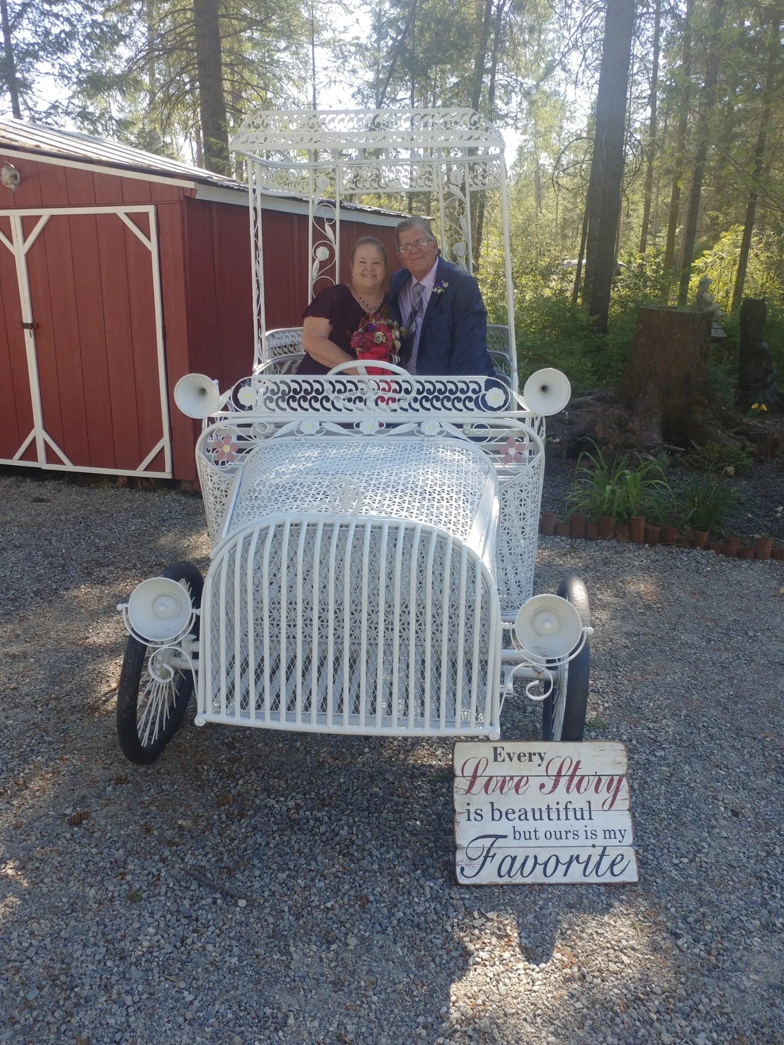 A couple dressed in wedding attire sitting in a white vintage carriage with decorative ironwork, outdoors with trees and a red shed in the background.
