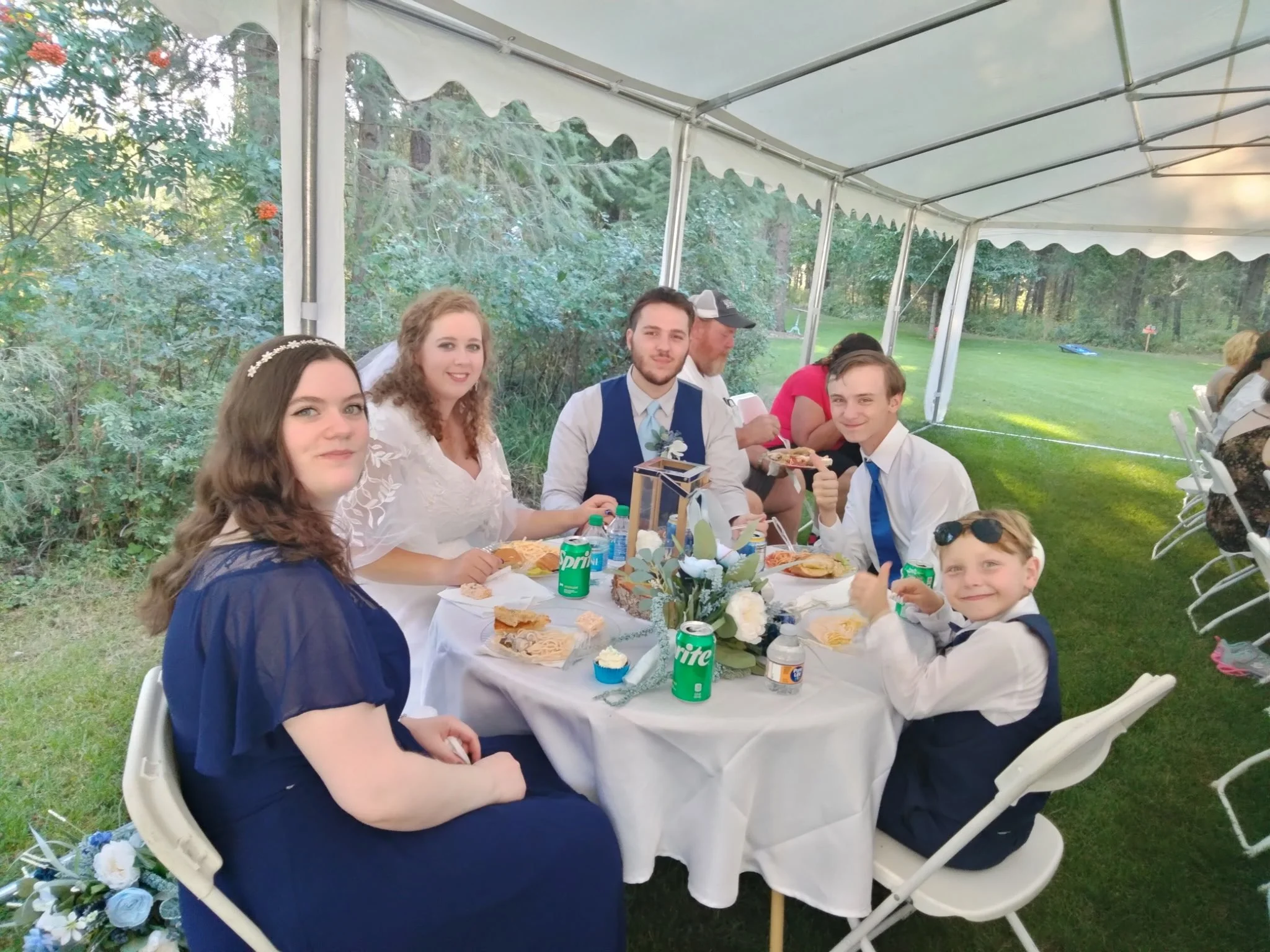 A family or group of people sitting at a table under a white canopy outdoors, smiling and having a meal. The table has a white tablecloth, floral centerpiece, and various food and drinks.