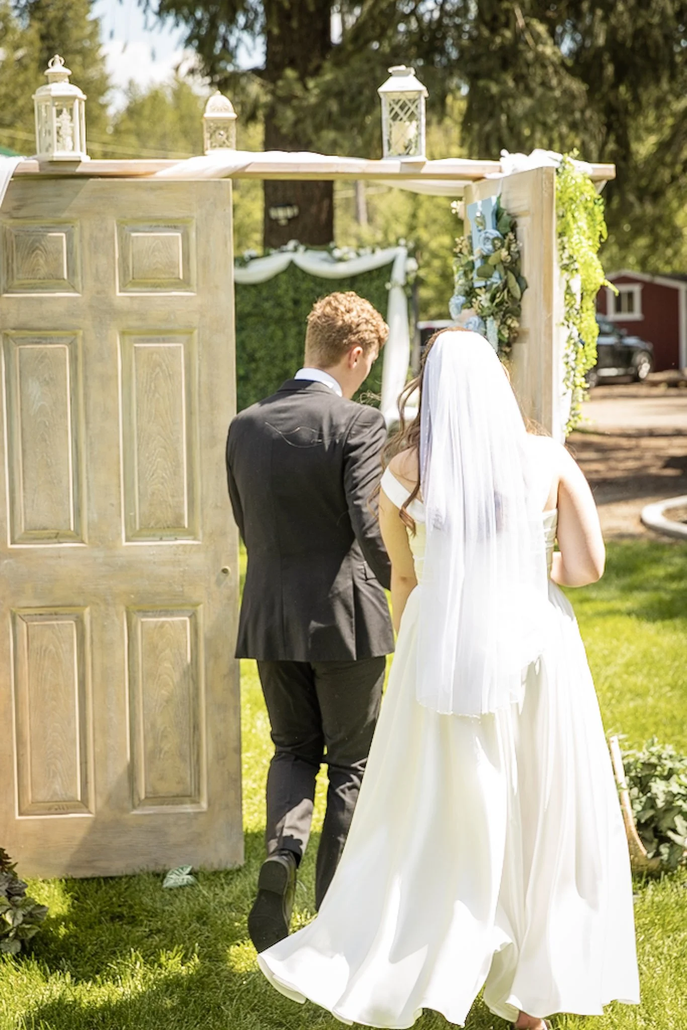 A bride and groom standing outdoors at a wedding ceremony under a decorative wooden arch decorated with flowers and greenery, with a backdrop of trees and a red barn.