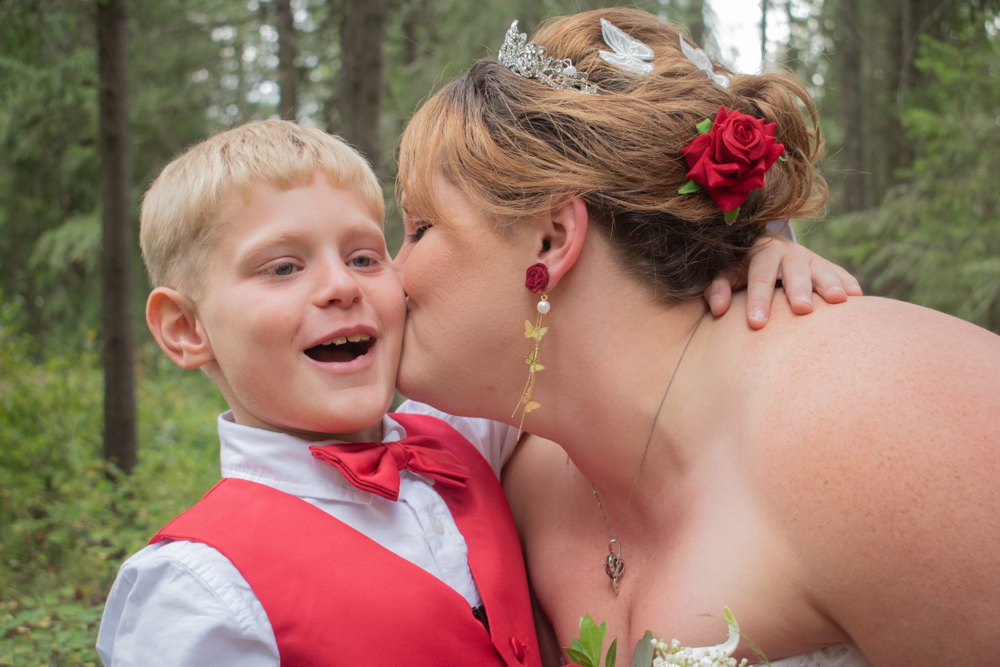 A young boy in a white shirt and red vest kissing a woman on the cheek in an outdoor forest setting.