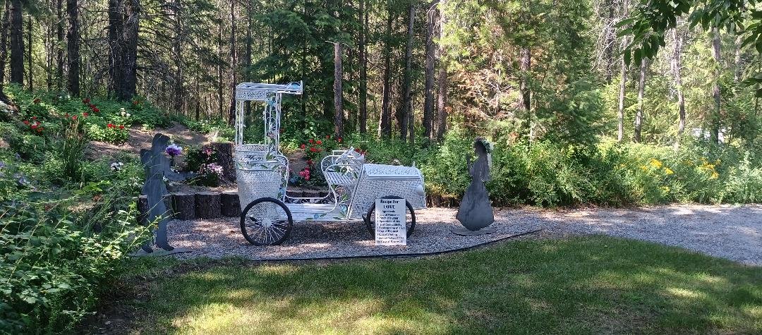 A roadside memorial with a white metal wheelchair sculpture, surrounded by flowers, trees, and greenery in a wooded area.