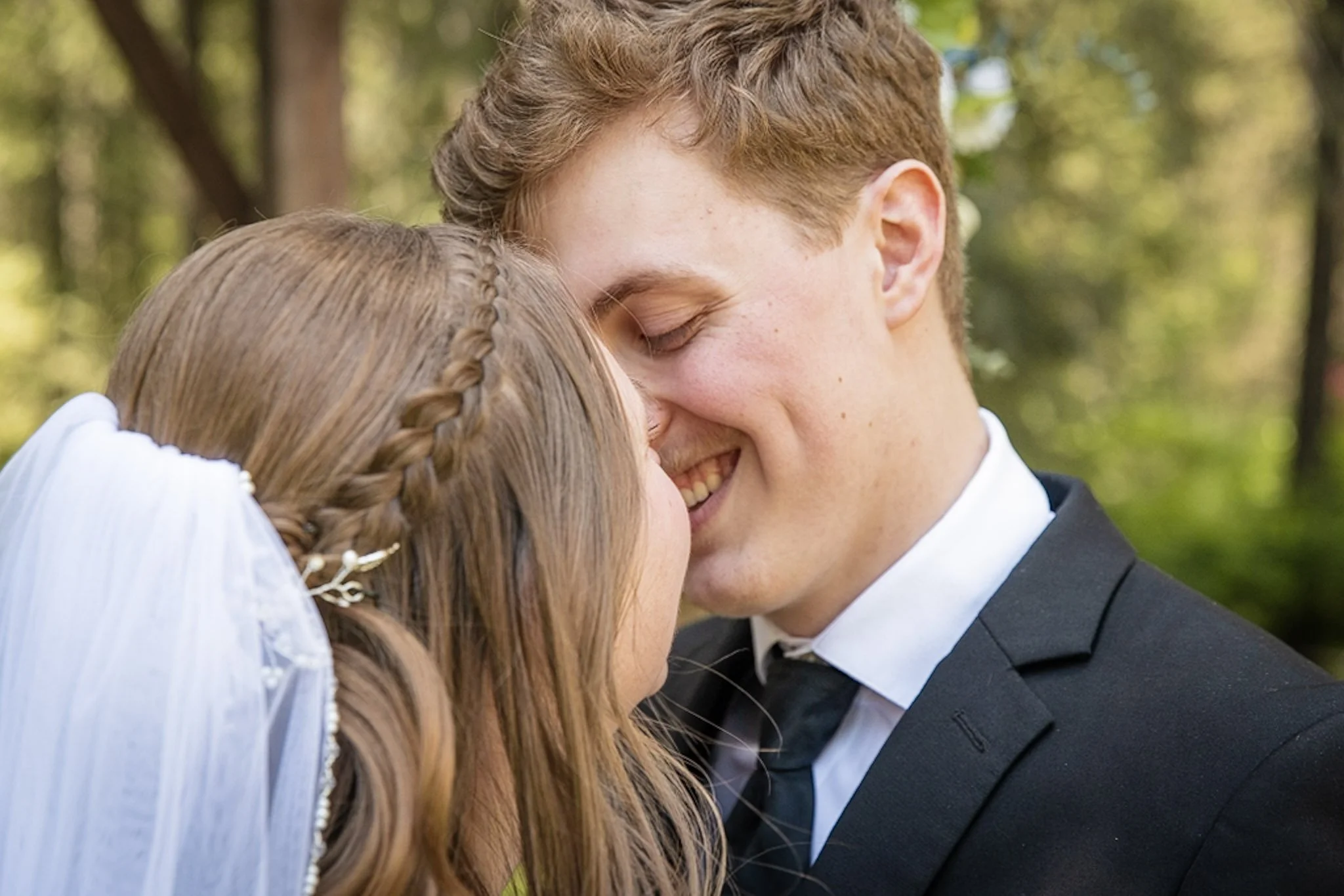 A young couple sharing an intimate moment outdoors, with both of their eyes closed, smiling, and touching foreheads.