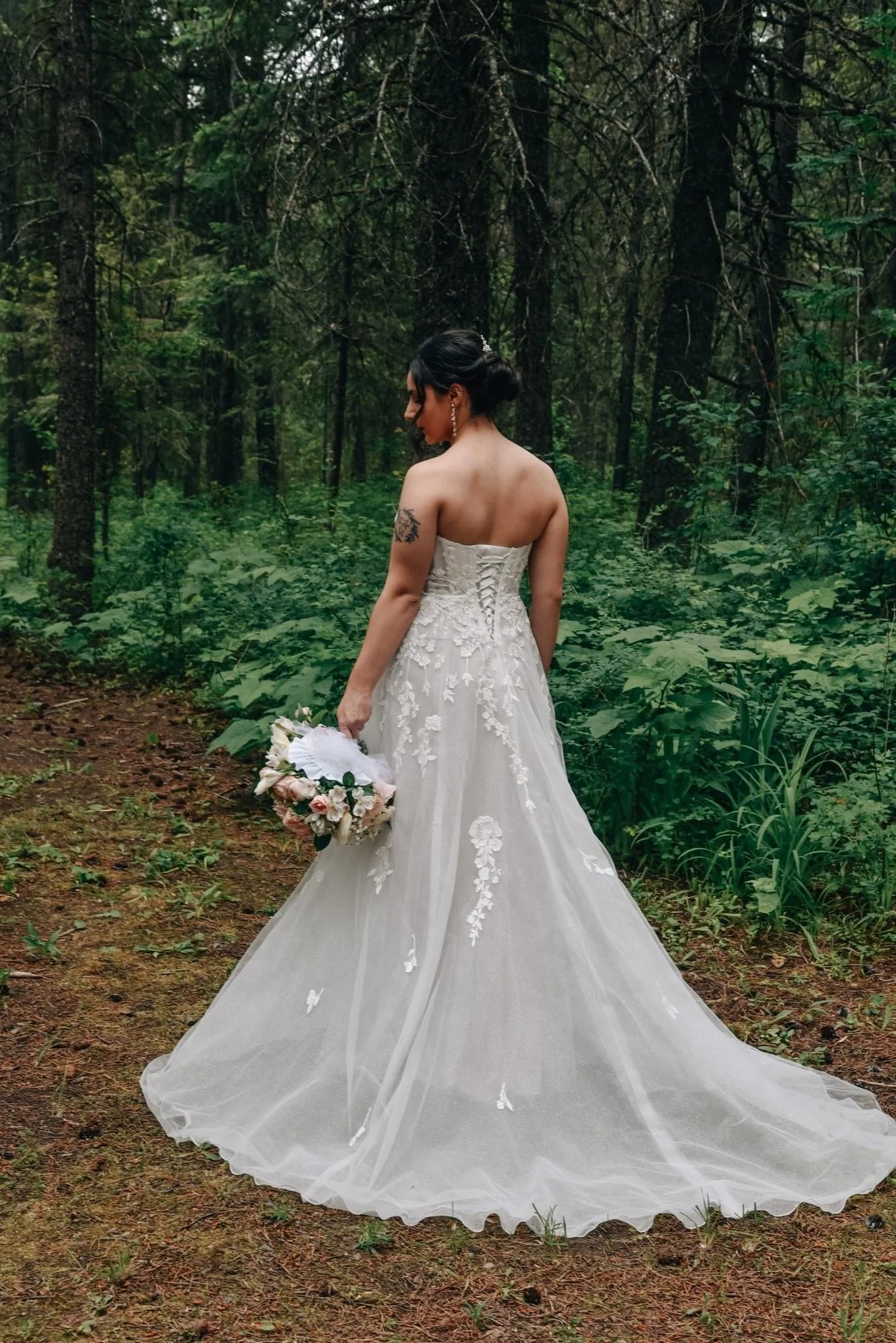 A bride in a strapless white wedding gown holding a bouquet in a lush green forest.
