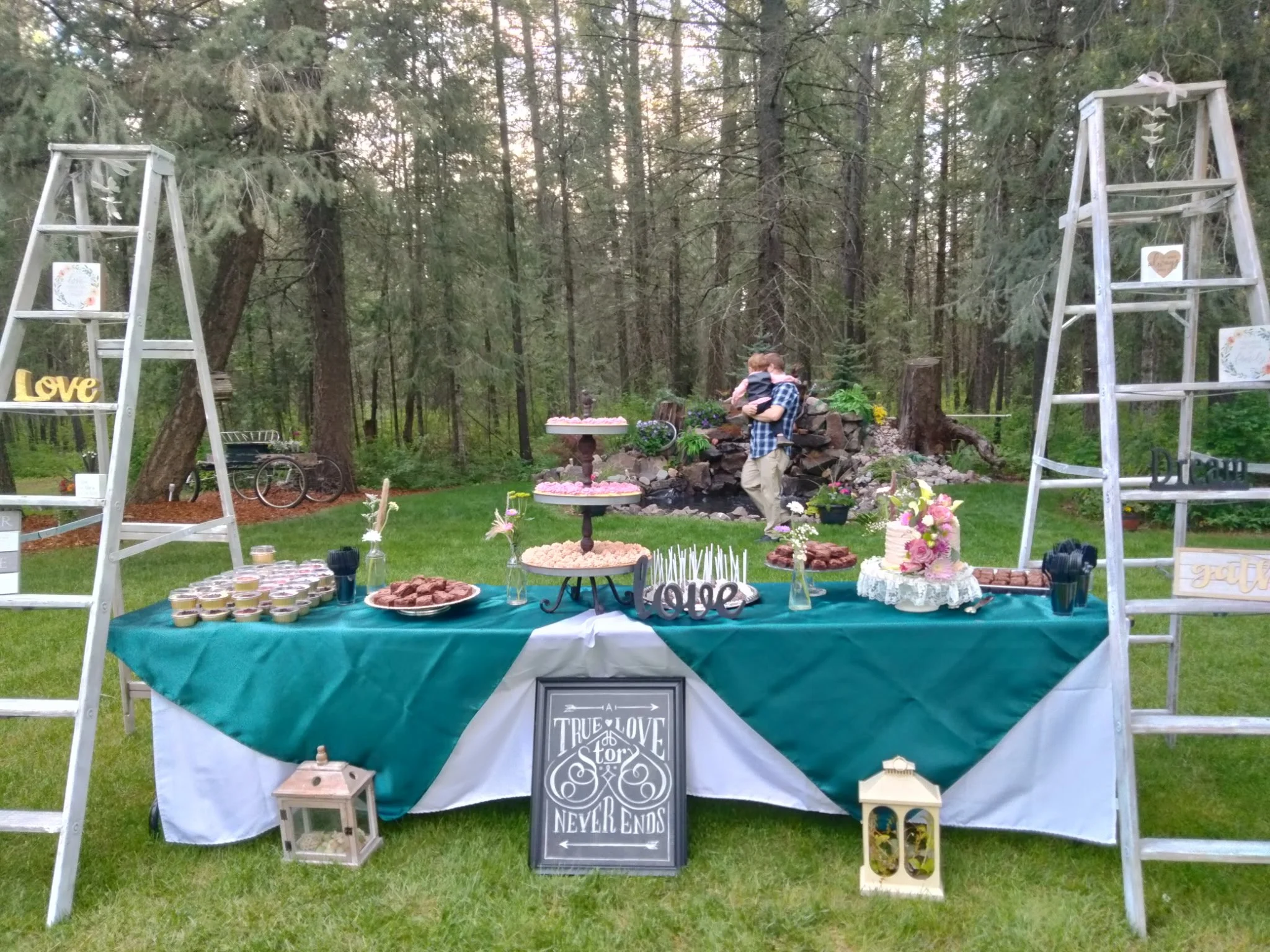 Outdoor celebration table decorated with teal and white tablecloth, cupcakes, cookies, flowers, and a cake, set in a forested area with trees in the background. Two white ladders with decorative signs and pictures flank the table. A man holds a child