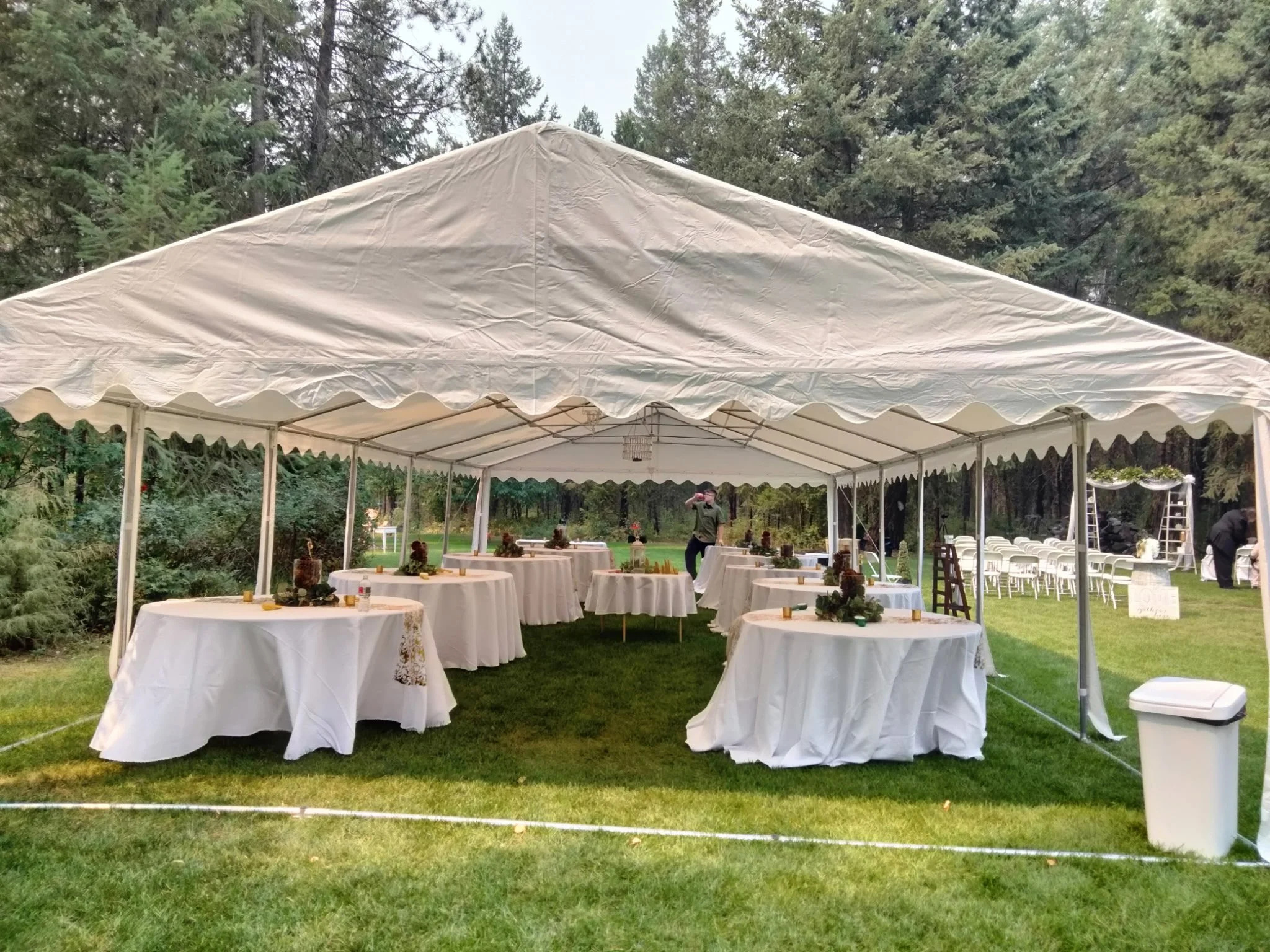 White event tent set up outdoors on grass with round tables covered in white tablecloths, decorated with greenery centerpieces. Additional chairs are visible in the background, and forest trees surround the area.