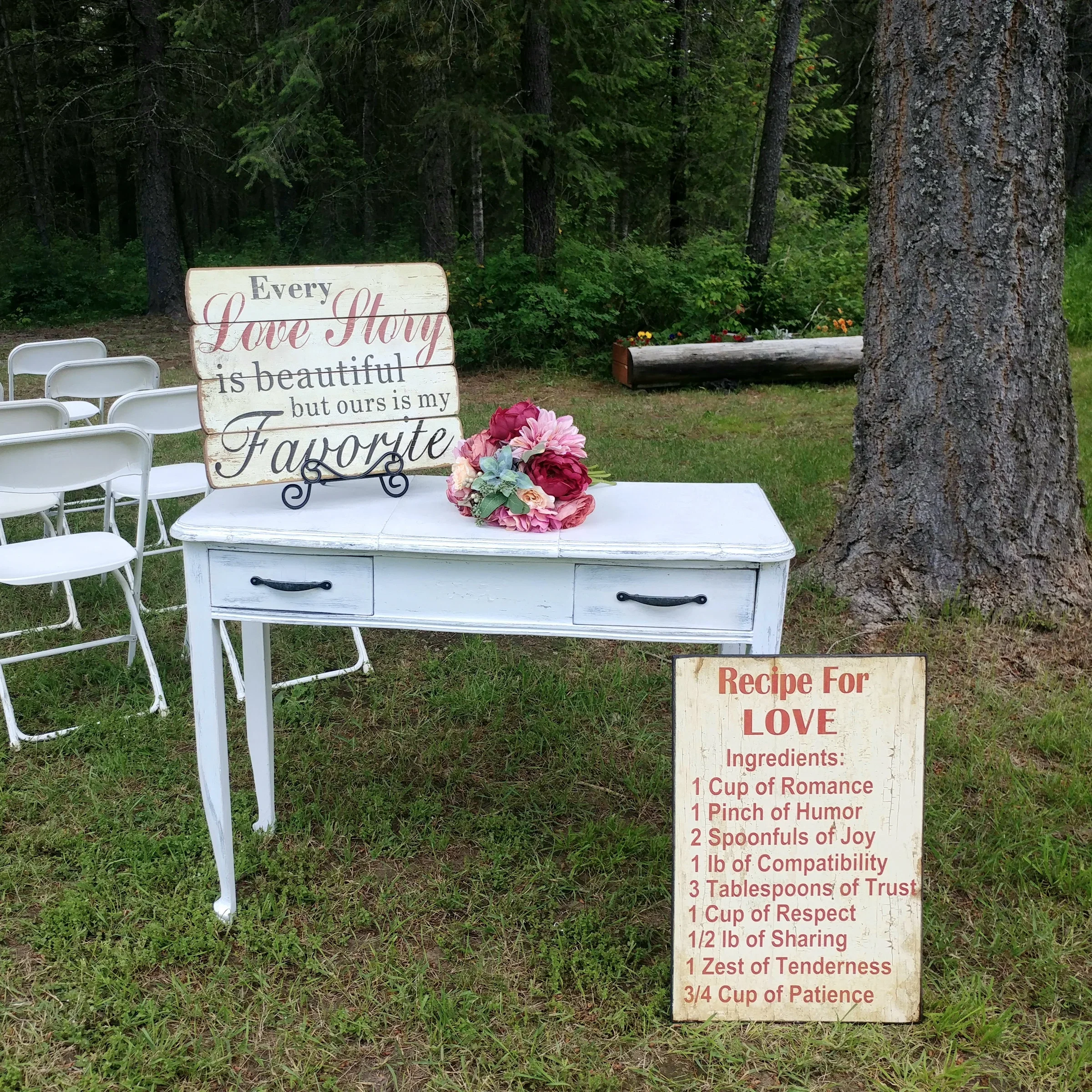 Outdoor setting with a white table and chairs, a decorative sign that reads "Every Love Story is beautiful but ours is my Favorite," a bouquet of pink and purple flowers, and a rustic wooden sign with a love recipe listing ingredients like romance, h