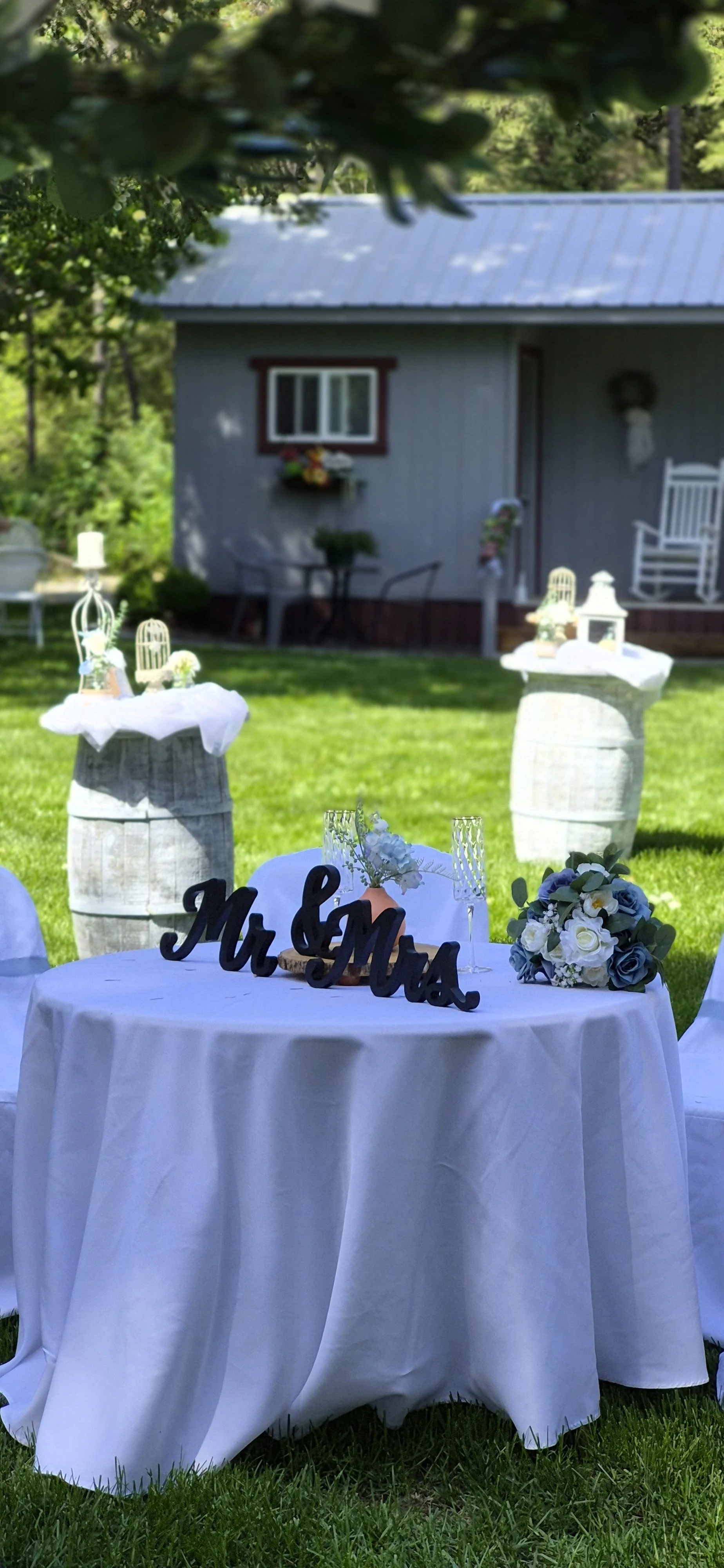 Outdoor wedding reception setup with a round table covered in a white tablecloth, decorated with 'Mr & Mrs' sign, floral arrangements, and white chairs. In the background, there are decorative barrels, lanterns, a house with a window, a rocking chair