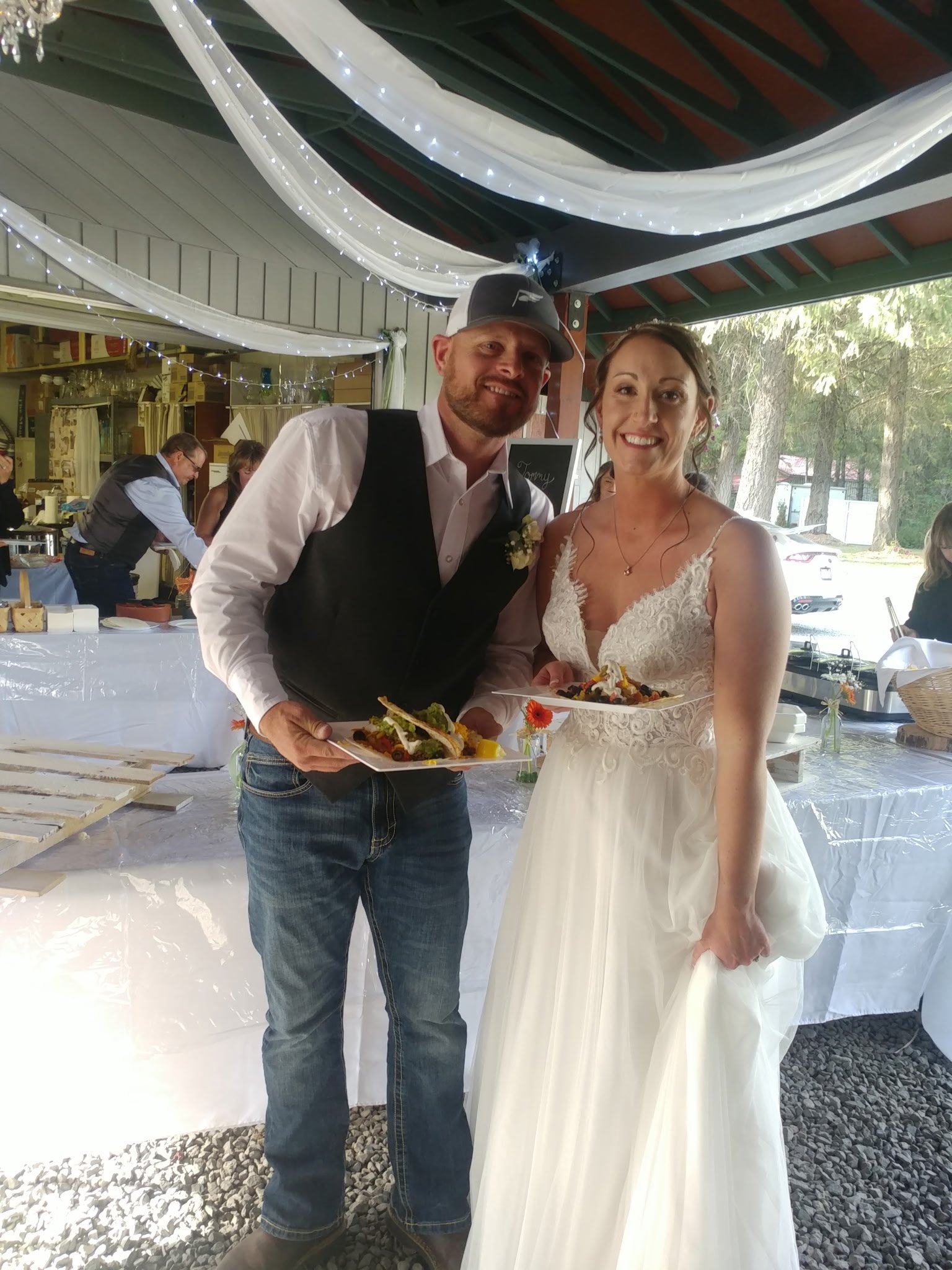 A bride and groom at a wedding reception, holding plates of food and smiling for the camera, indoors with white draped fabric and string lights overhead.