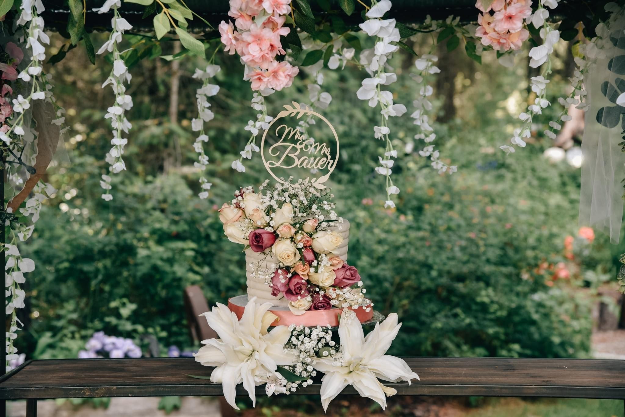 A wedding cake decorated with white and purple flowers, including lilies and roses, set on a rustic wooden table outdoors. Hanging floral arrangements frame the scene with a sign reading 'Mom Mom Bauer' at the top.