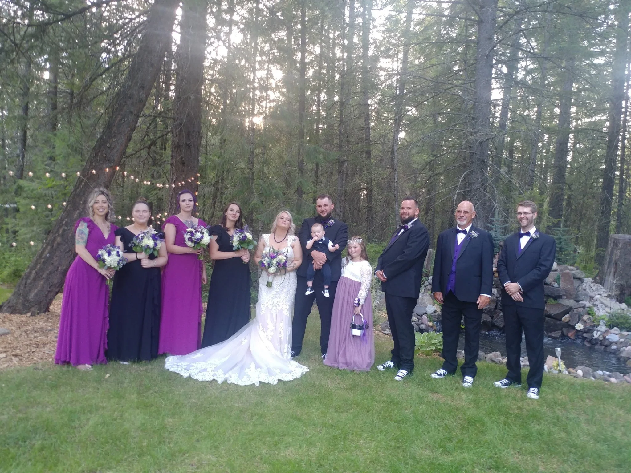 A wedding party standing outdoors in a wooded area with string lights, including bridesmaids in purple and black dresses, the bride in a white gown, groomsmen in black tuxedos with purple vests, and children, during daylight.
