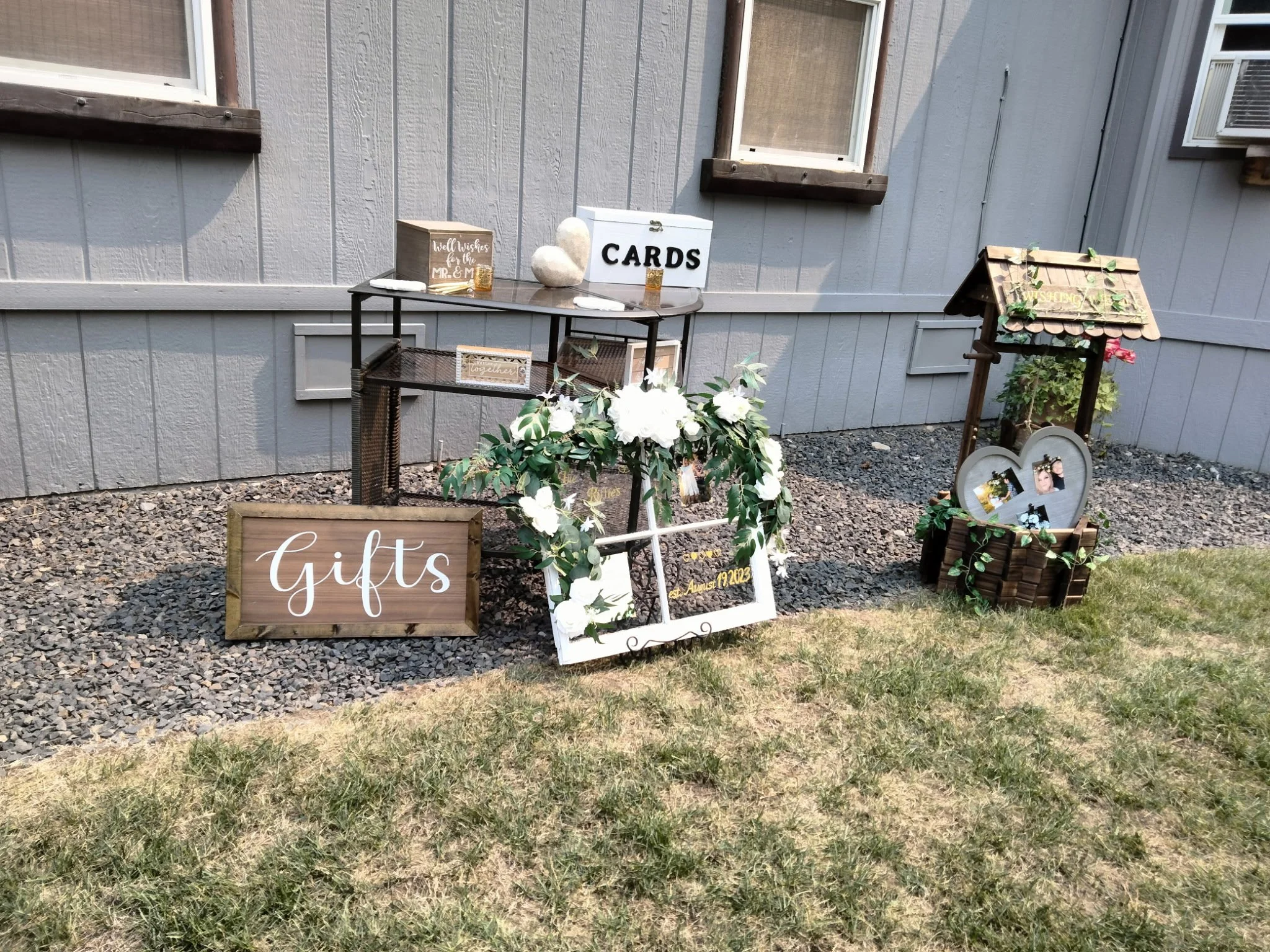 Outdoor display of gifts and cards setup with a wooden sign labeled "Gifts," a black table holding cards, a white frame with flowers, a large rock decoration, and a small wooden wishing well with greenery and framed photographs inside.
