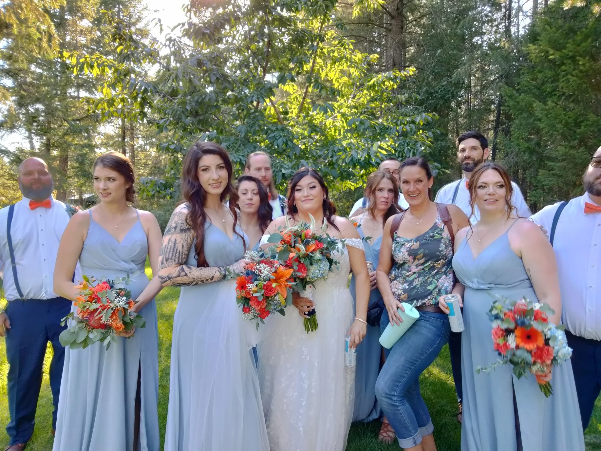 A group of people at a wedding outdoors, with women in blue dresses and men in white shirts with suspenders, some holding bouquets, standing on grass among trees.