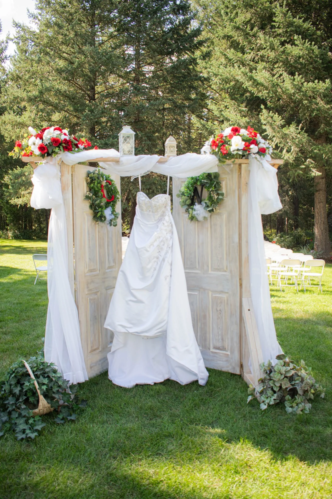 Outdoor wedding setup with a white wedding dress hanging on a makeshift wooden structure decorated with flowers, lanterns, and greenery, surrounded by chairs and trees.