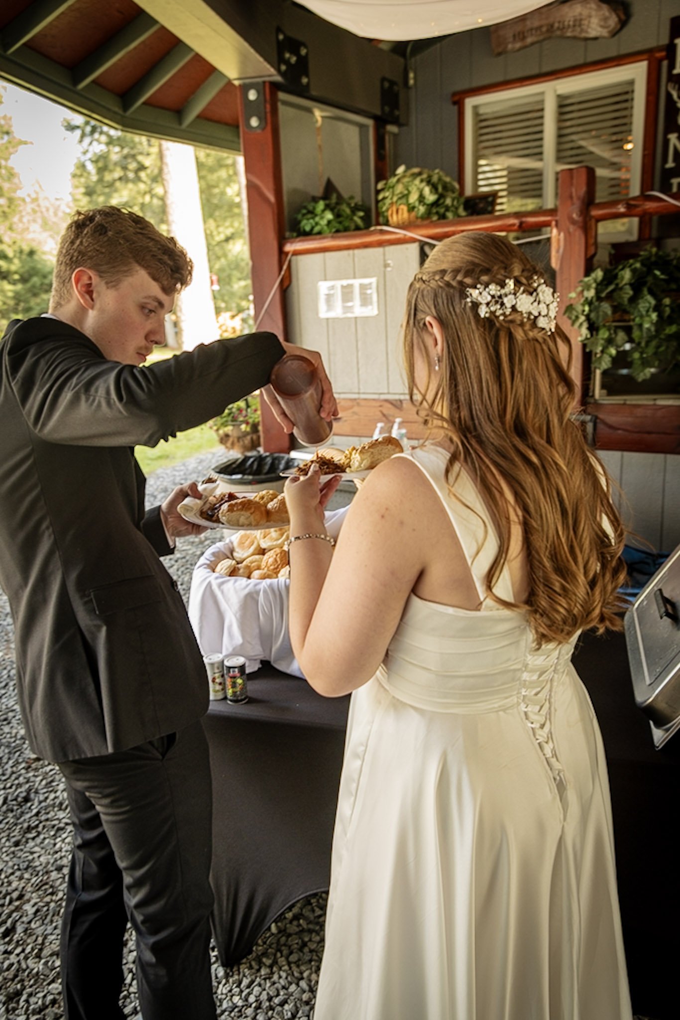A bride in a white wedding dress with a floral hair accessory is receiving food from a groom wearing a black suit at an outdoor wedding reception.