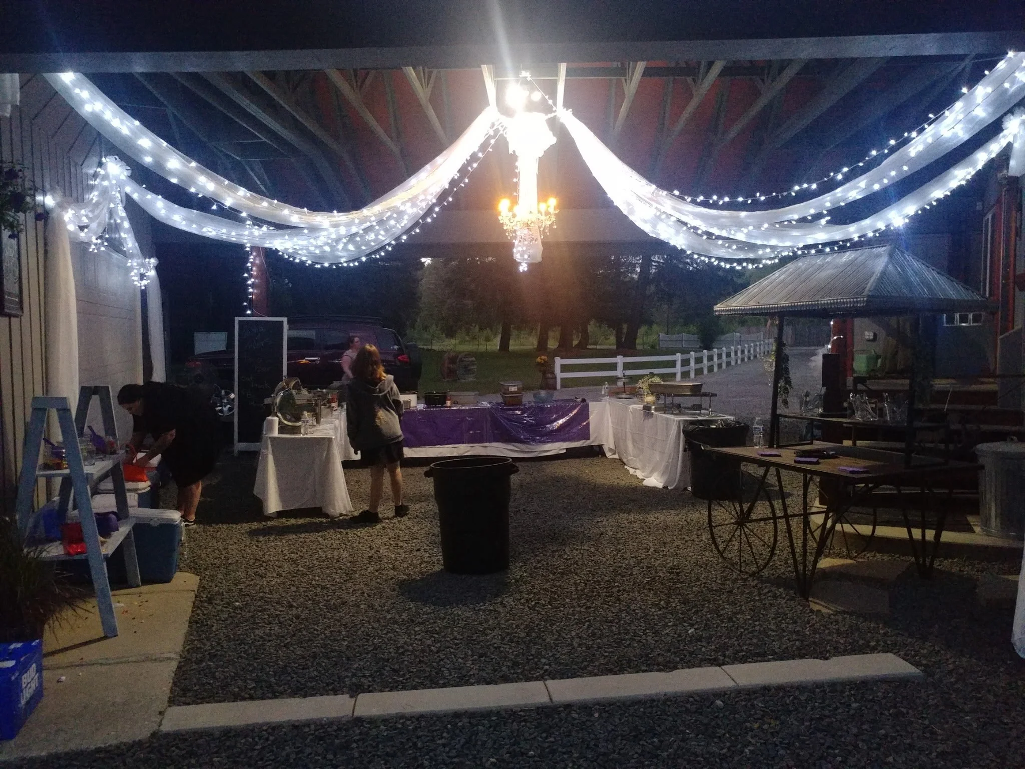 Outdoor evening setup with string lights and a chandelier, tables with white and purple tablecloths, a small gazebo, and people preparing for an event.