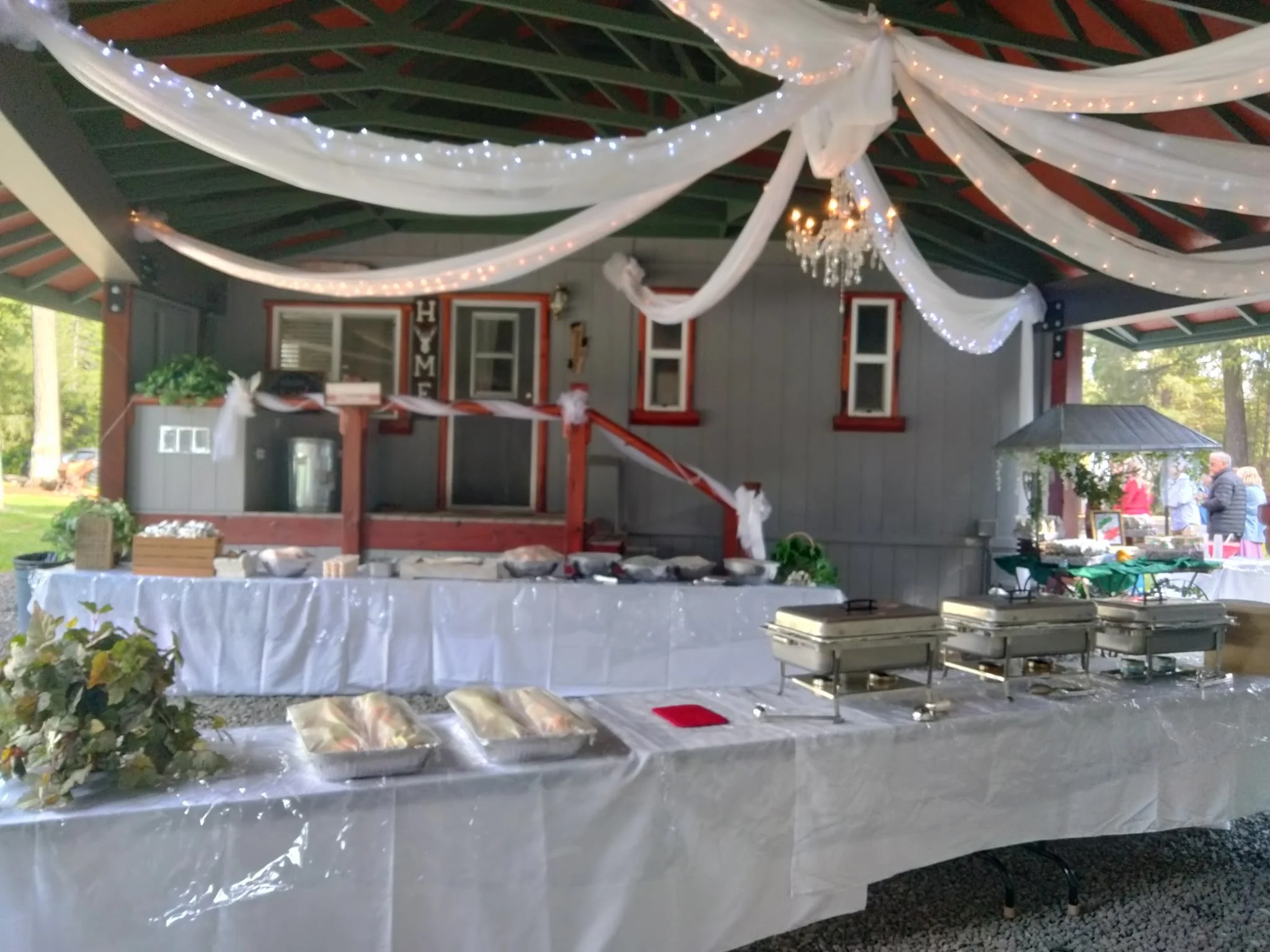 Buffet table with chafing dishes, wrapped in white tablecloth, decorated with white drapery and string lights; house with themed decorations in the background.