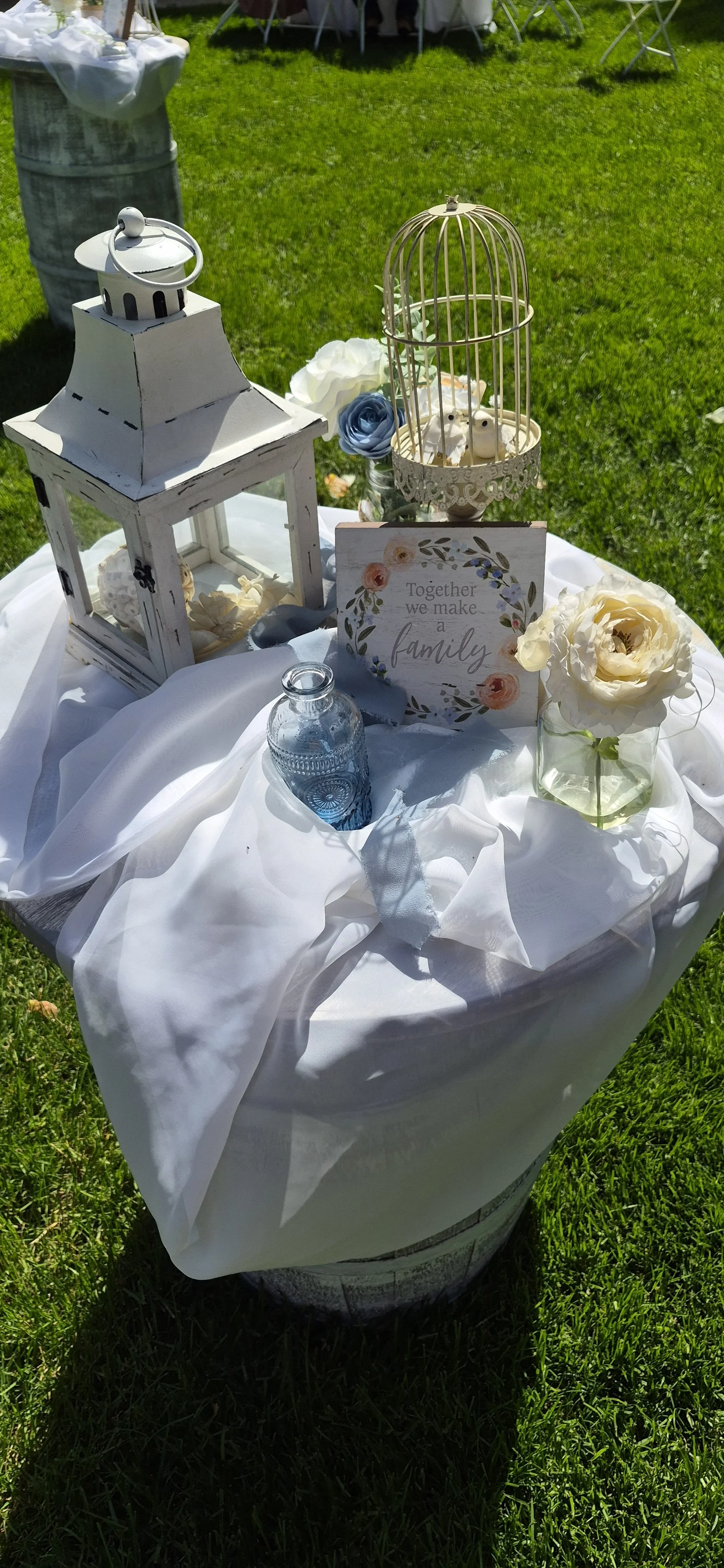 Decorative table setup with lanterns, a birdcage, flowers, a glass jar, and a sign that says, 'Together we make a family' outdoors on grass.
