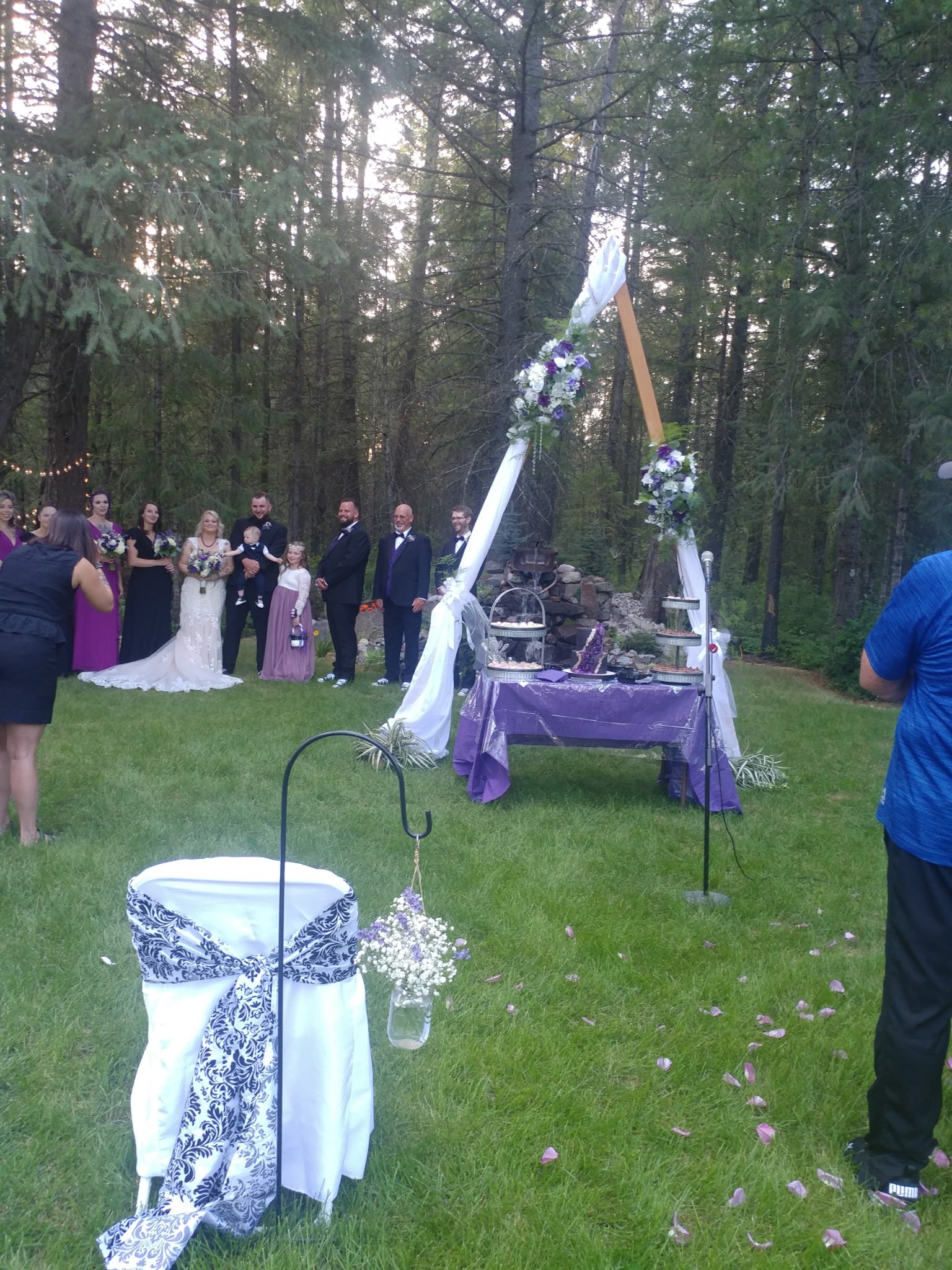 A wedding ceremony in a wooded outdoor setting with a decorated altar, a bride and groom, and wedding party members.