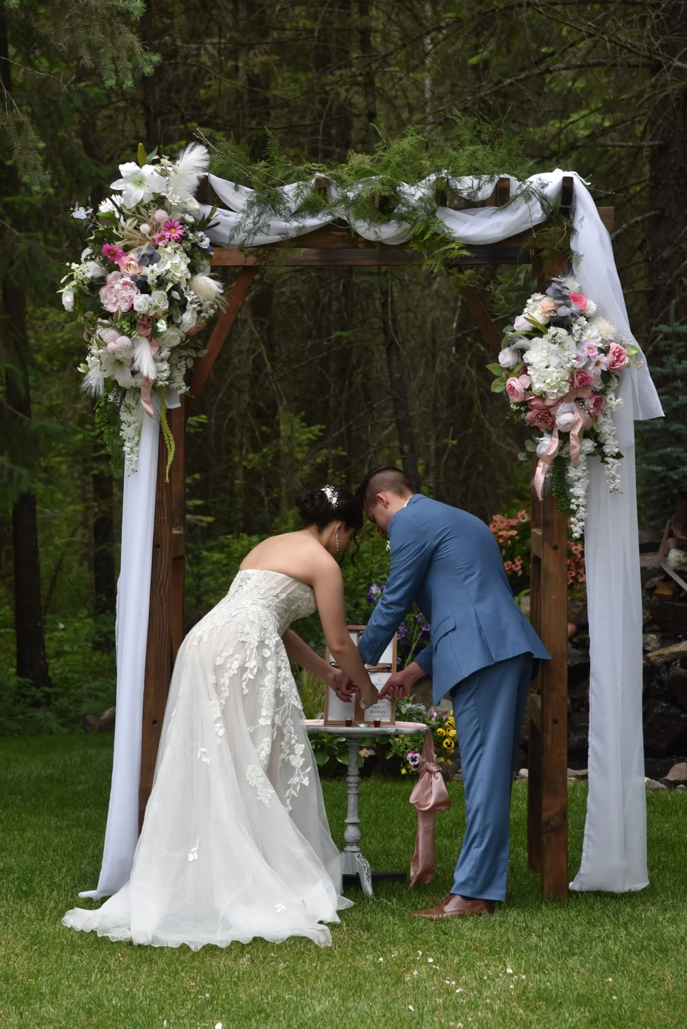 A bride and groom standing under a decorated wedding arch with flowers and greenery in an outdoor setting, exchanging rings.