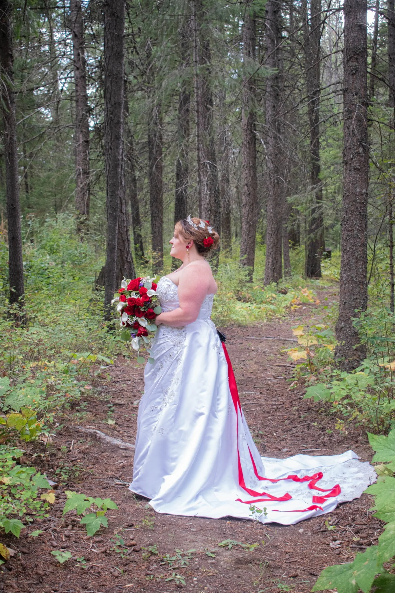 A bride standing on a forest trail, wearing a white wedding dress with red accents, holding a bouquet of red and white flowers, surrounded by tall trees and greenery.